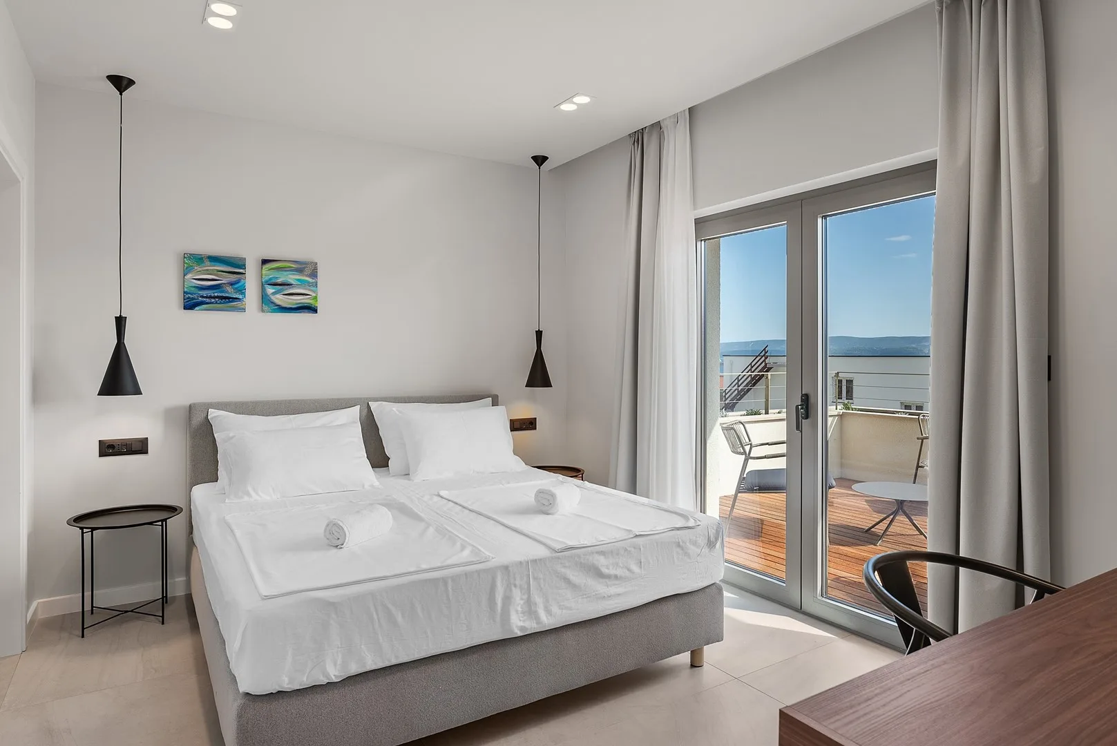 A bright bedroom with a neatly made bed beside glass doors opening onto a balcony.