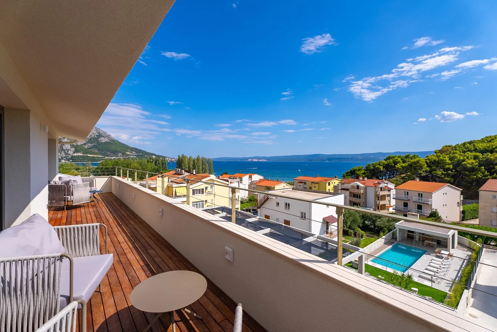 A balcony overlooks nearby buildings, a swimming pool, and the sea.