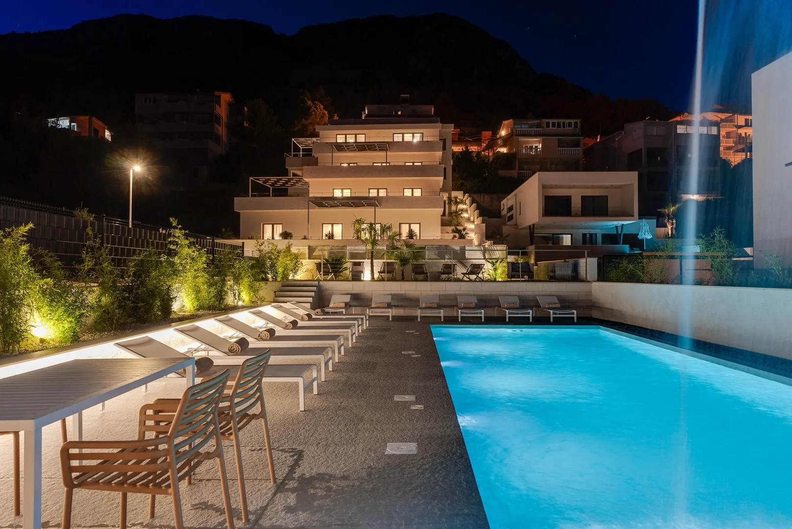 An illuminated outdoor pool beside a terrace seating area at night.