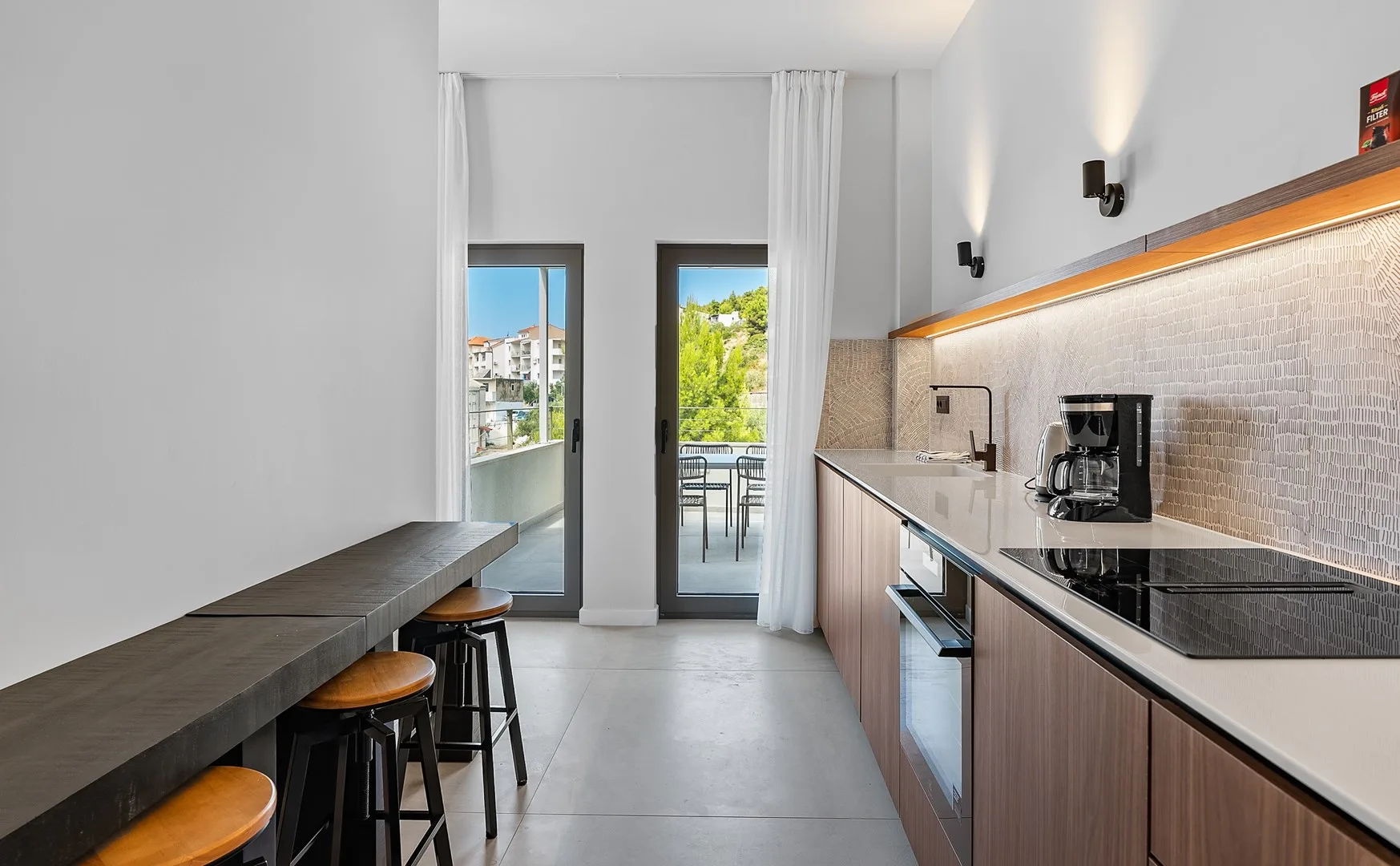 A modern kitchen with a long counter, cooktop, and glass doors opening to a balcony.