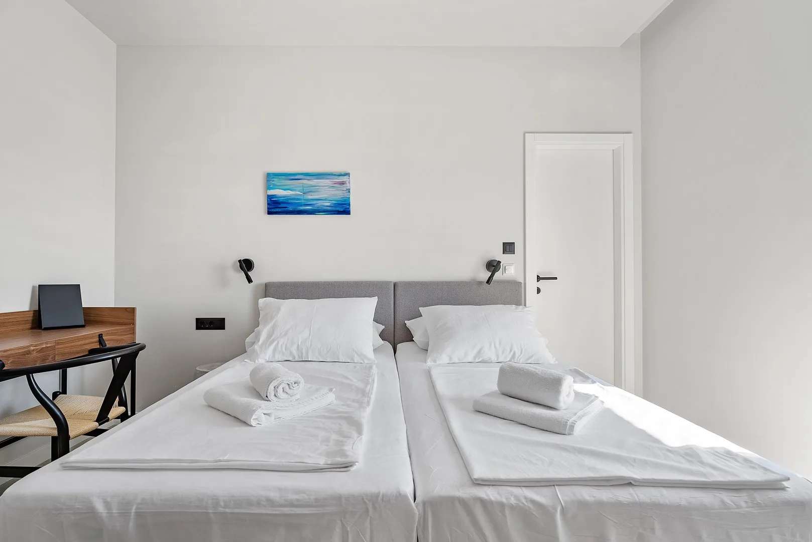 A bright bedroom with two beds, white linens, and a small wall painting.