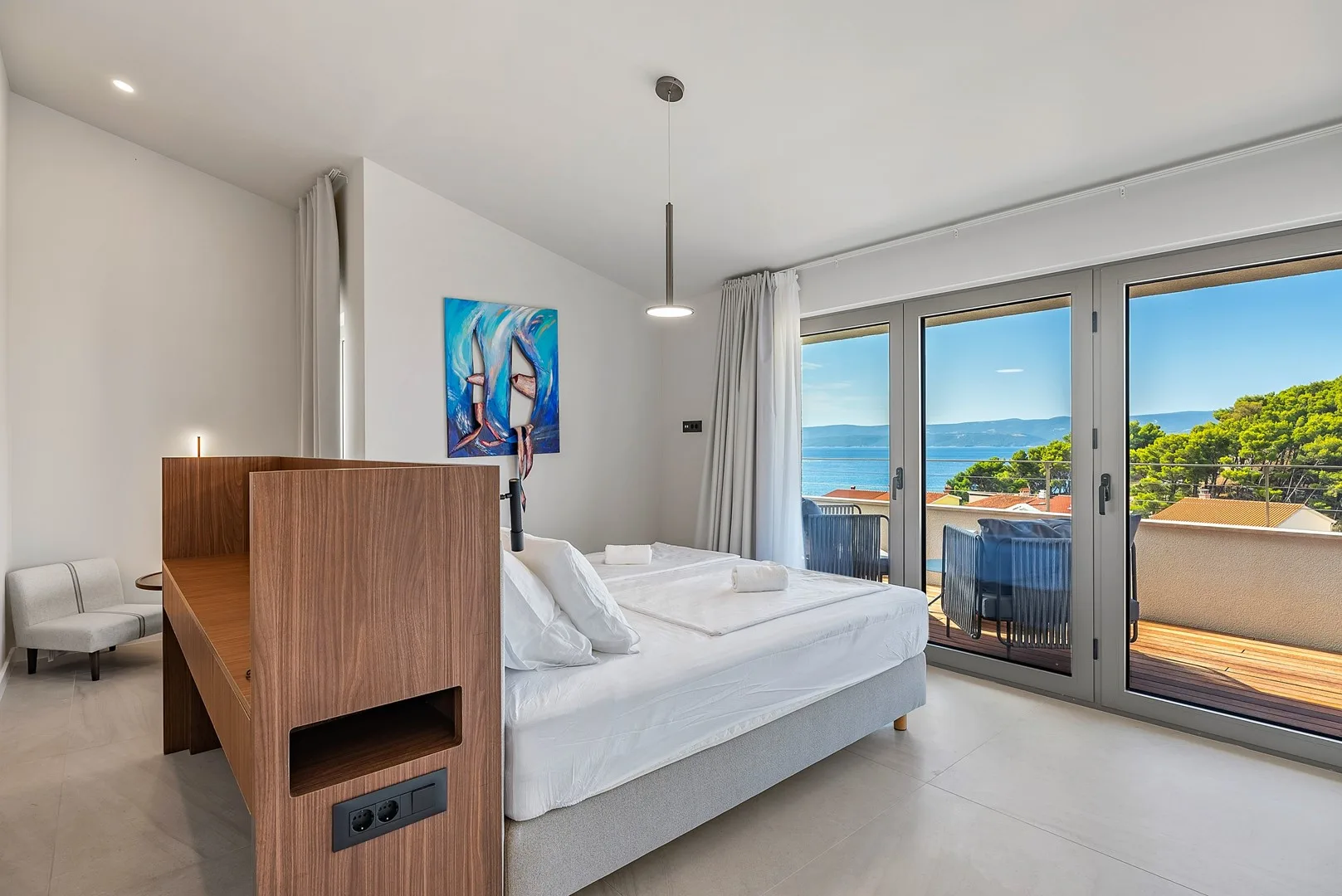A bright bedroom with a bed beside large glass doors overlooking the sea.