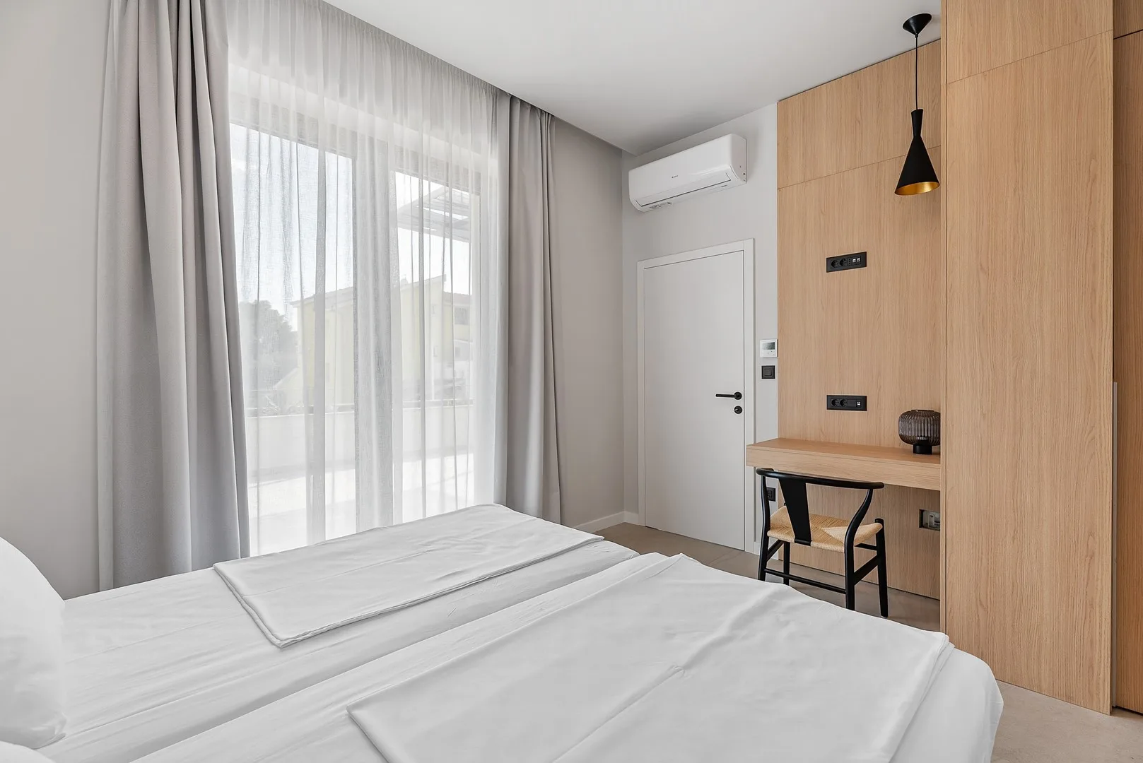 A bedroom with a neatly made bed beside a large curtained window.