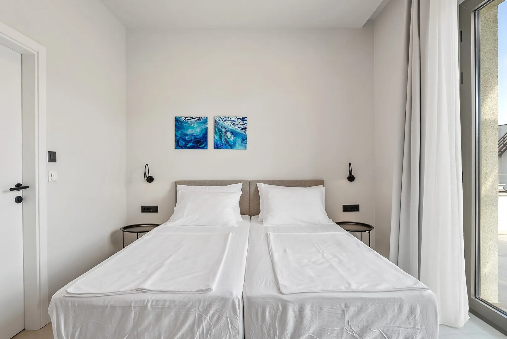 A bright bedroom with two beds beneath blue wall art.