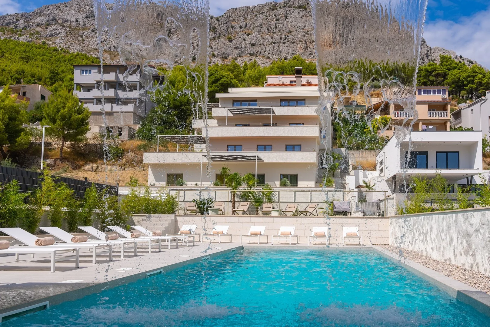 An outdoor swimming pool is surrounded by sun loungers, greenery, and hillside buildings.