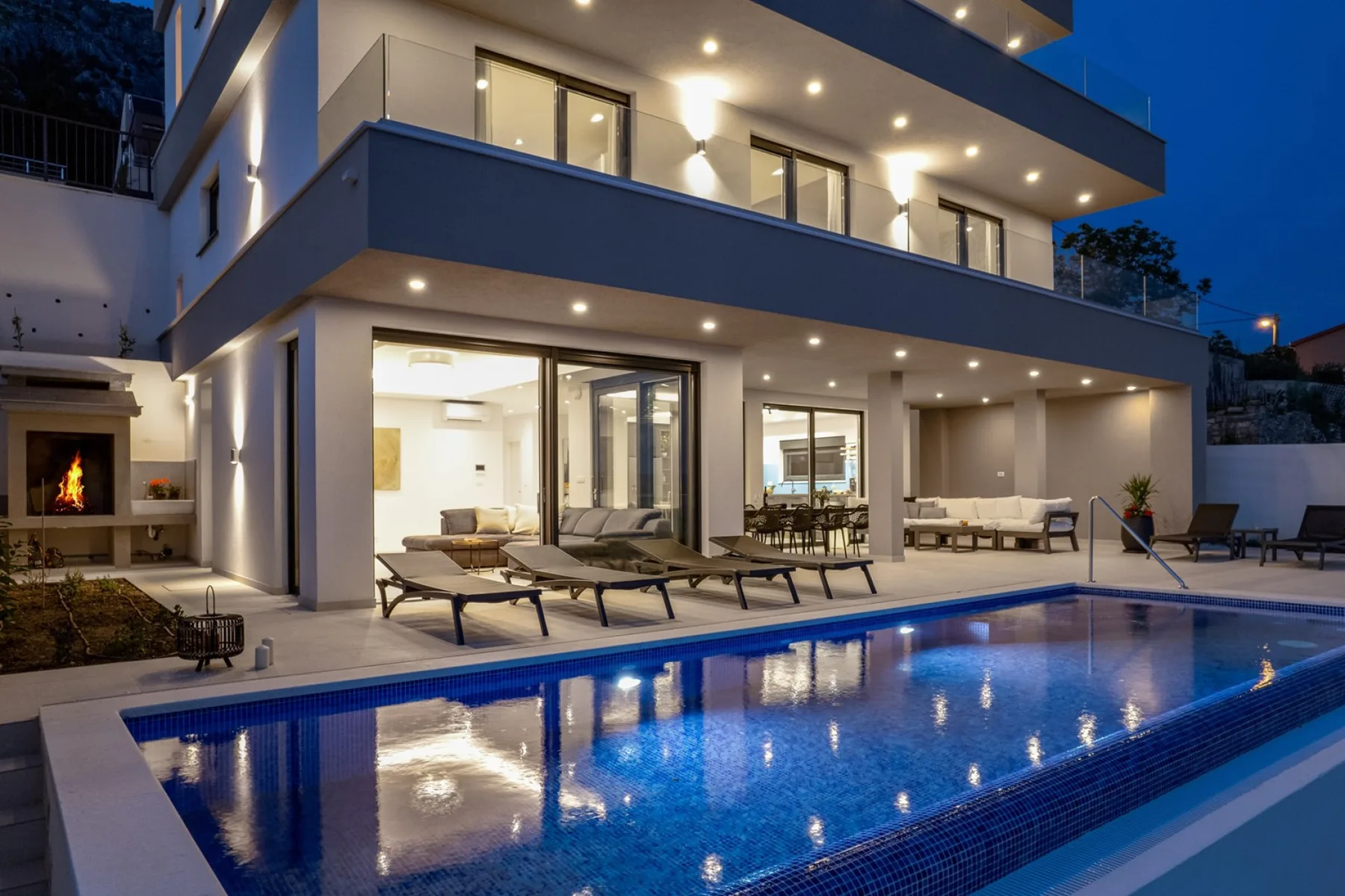 A modern multi-story house overlooks an illuminated swimming pool and outdoor seating at dusk.