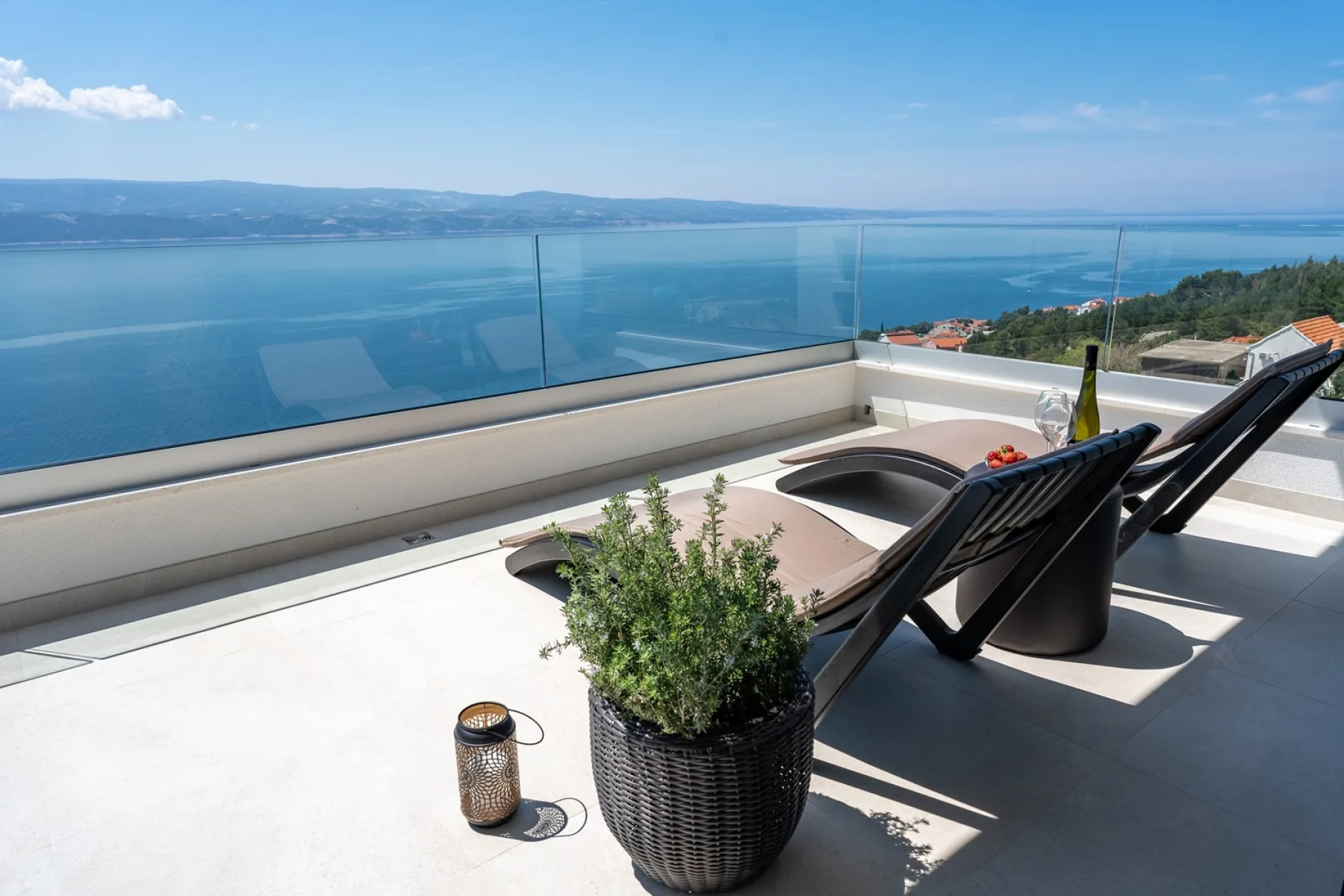 A sunlit terrace overlooks a lake, with lounge seating and potted greenery.