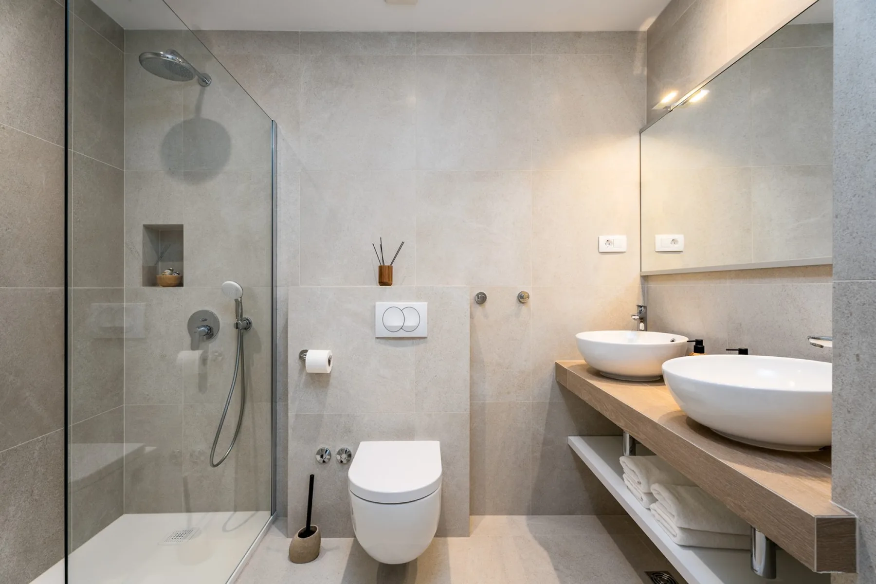 A modern tiled bathroom with a glass shower, toilet, and double sinks.