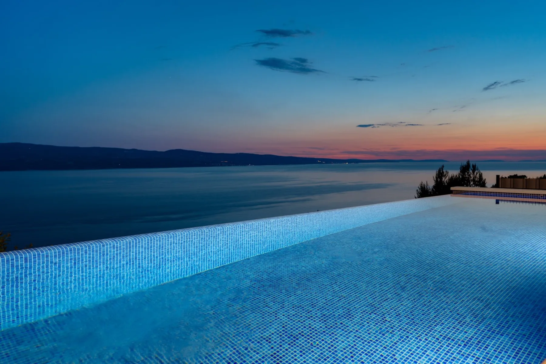An infinity pool overlooks calm water beneath a colorful twilight sky.
