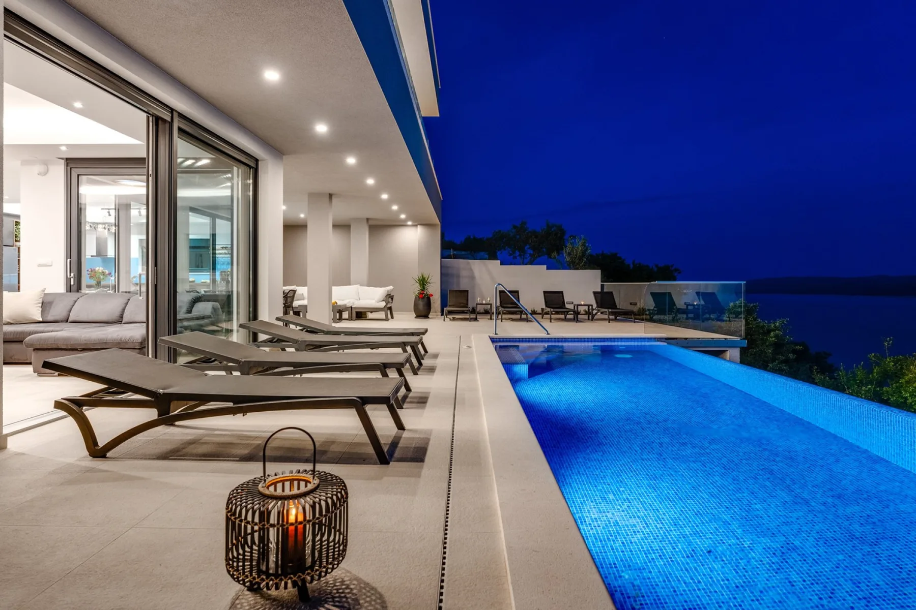 A modern outdoor pool beside a brightly lit patio with lounge seating.