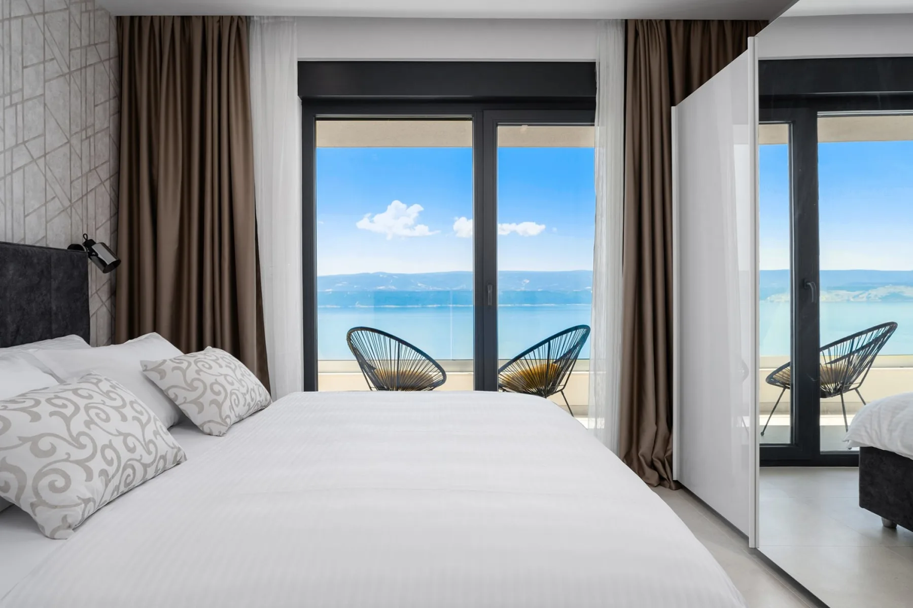 A bedroom with a white bed and a view of the sea through large windows.