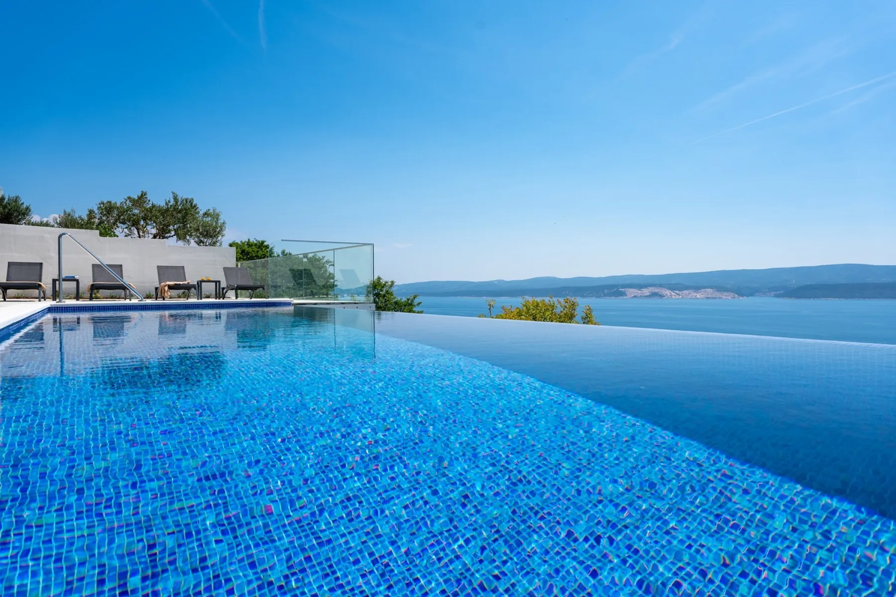 A blue swimming pool overlooks the sea, with lounge seating beside it.