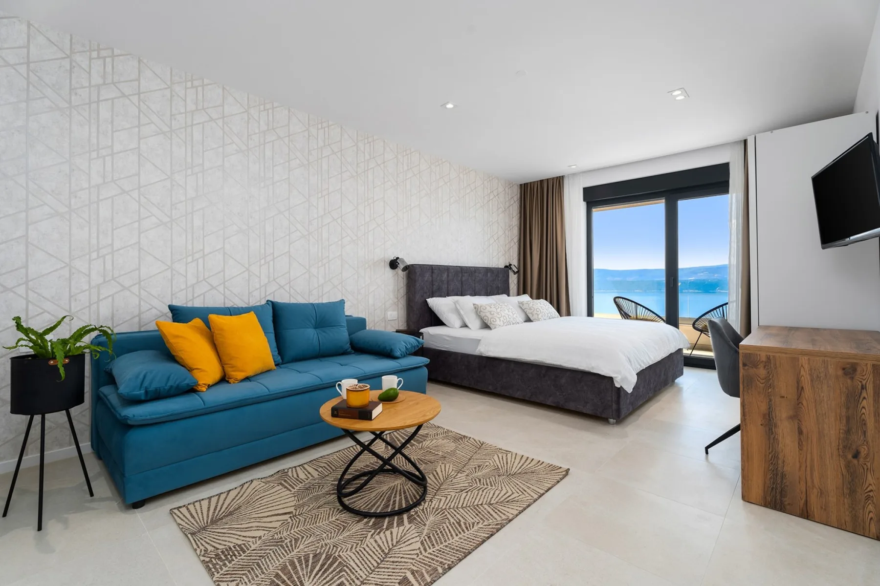 A bedroom sitting area with a teal sofa and a bed beside a balcony window.