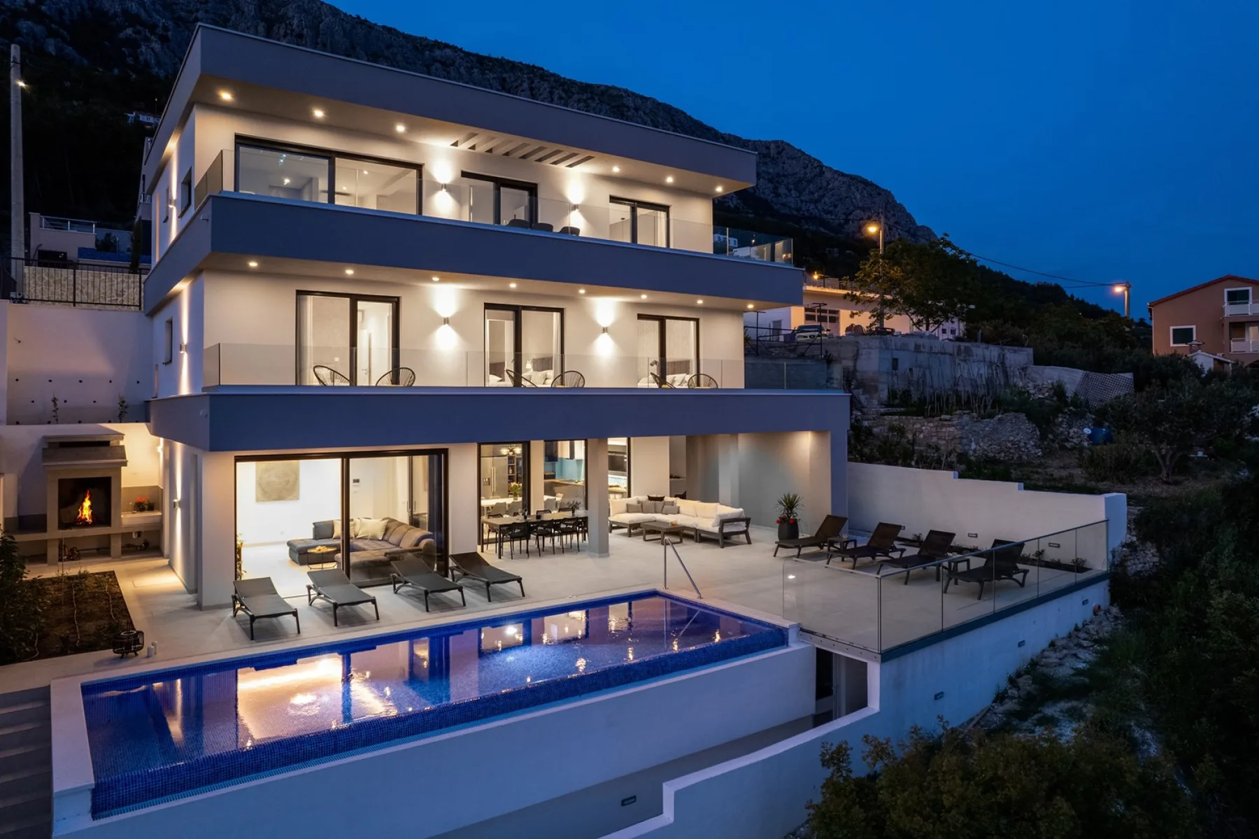 A modern multi-level house overlooks an outdoor pool and patio at dusk.