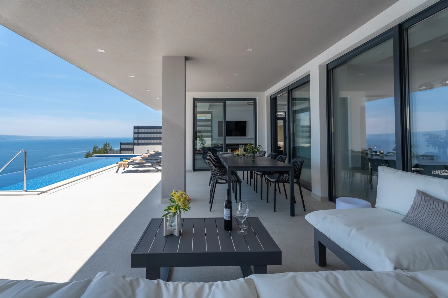 A covered terrace with outdoor seating overlooks a swimming pool and the sea.