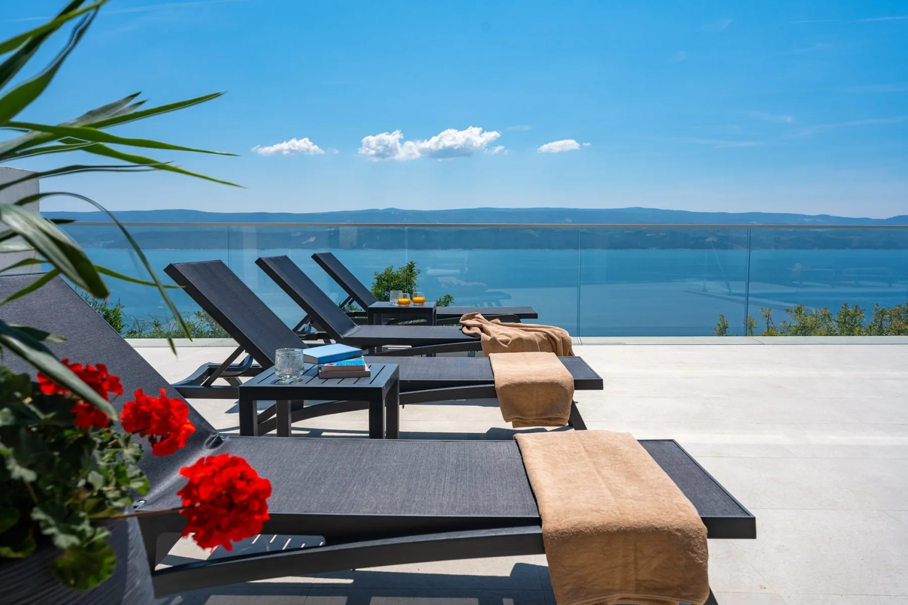 A waterfront terrace has dark lounge seating, tan towels, and a glass railing.