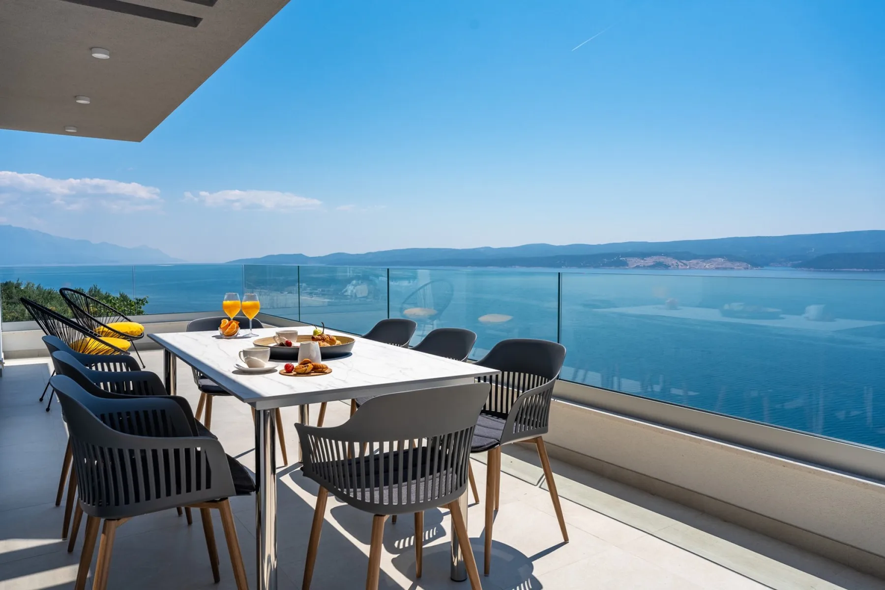 A dining table and seating on a terrace overlook the water and distant hills.