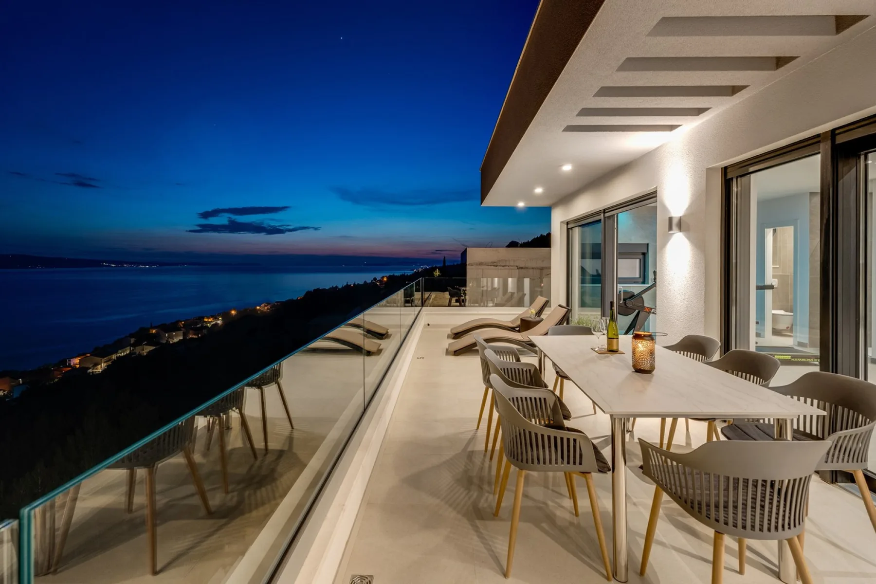 A seaside terrace with outdoor dining seating overlooks the water at dusk.