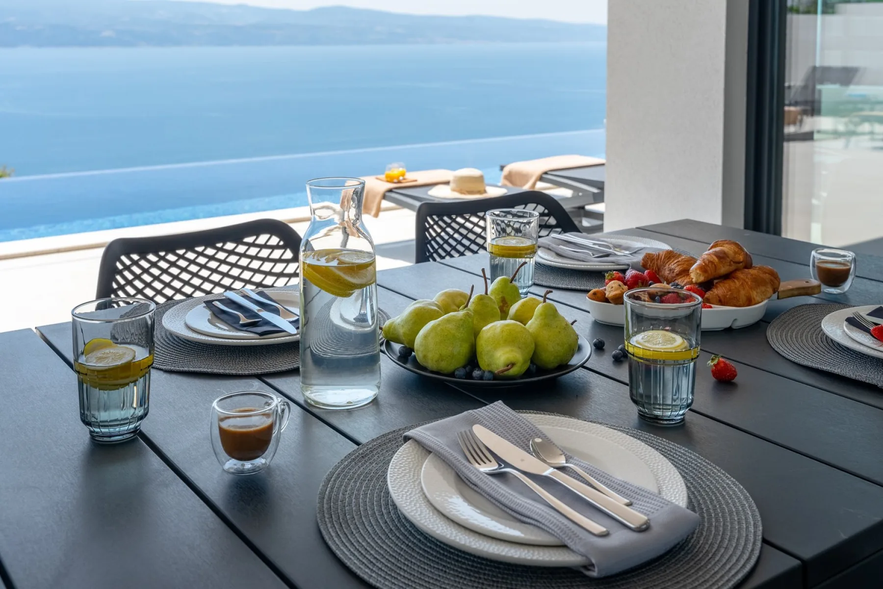 A dining table set with fruit and breakfast beside a pool overlooking the sea.
