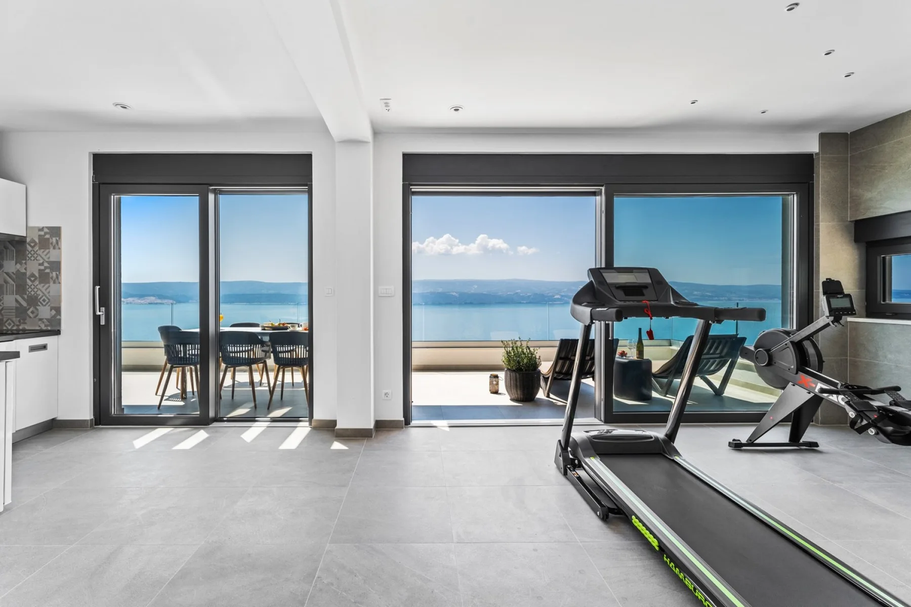 A bright indoor exercise area with treadmills beside large windows overlooking the sea.