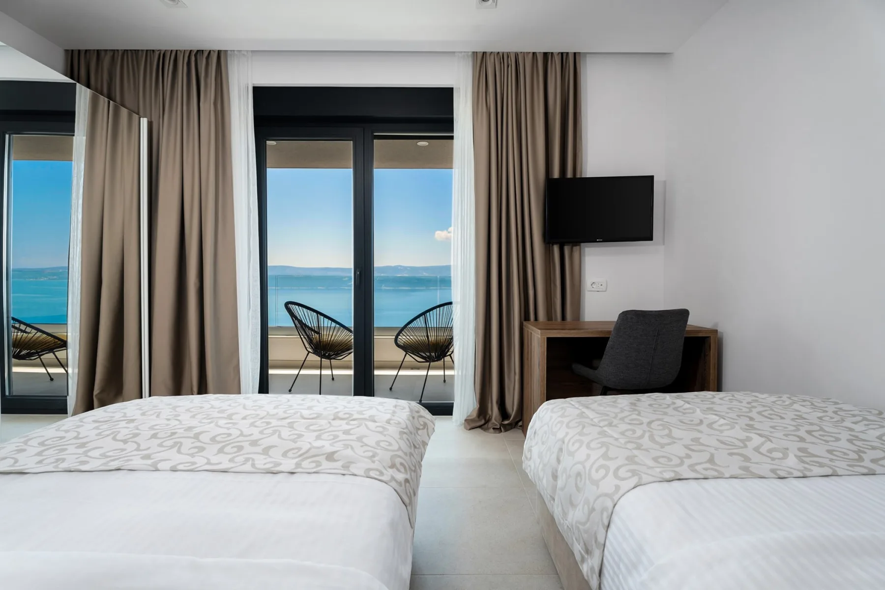 A bedroom with two beds, a desk, and glass doors overlooking the sea.