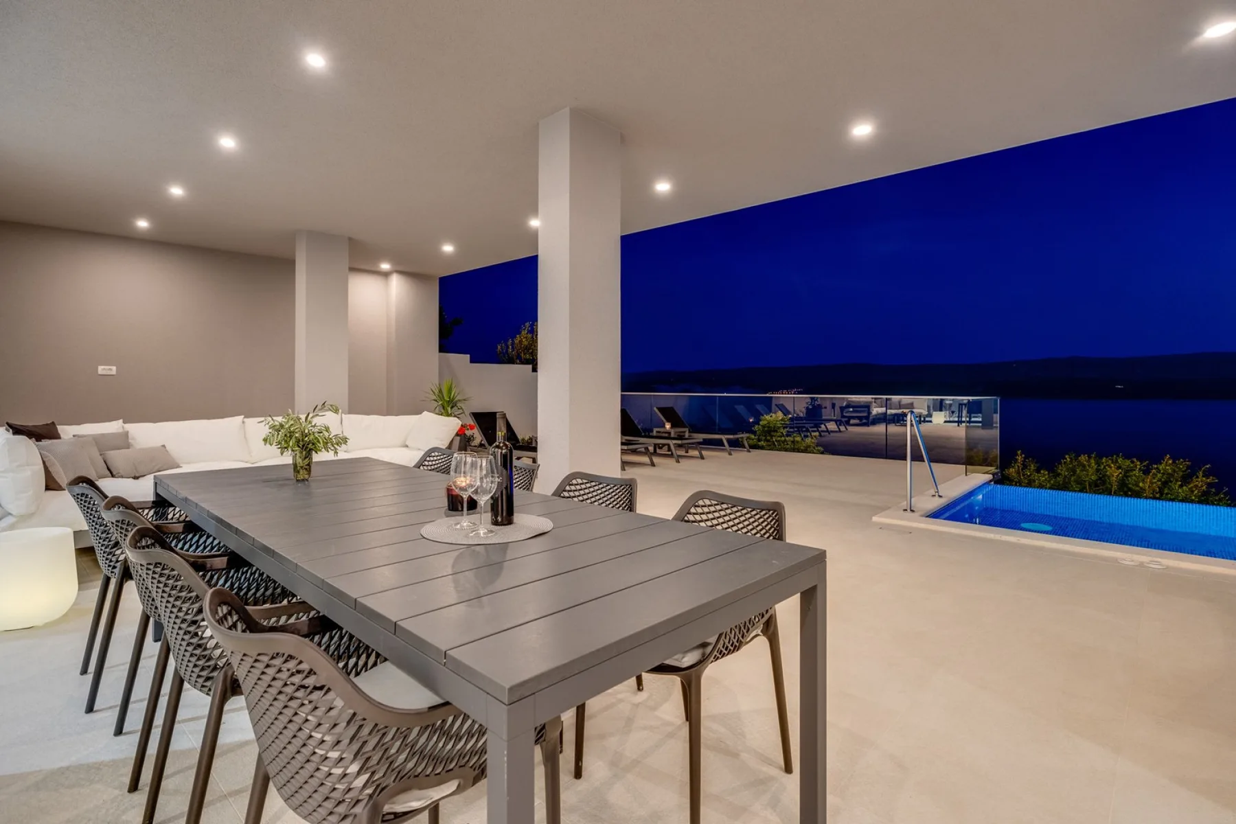 An outdoor dining area with a long table overlooks a lit swimming pool at night.