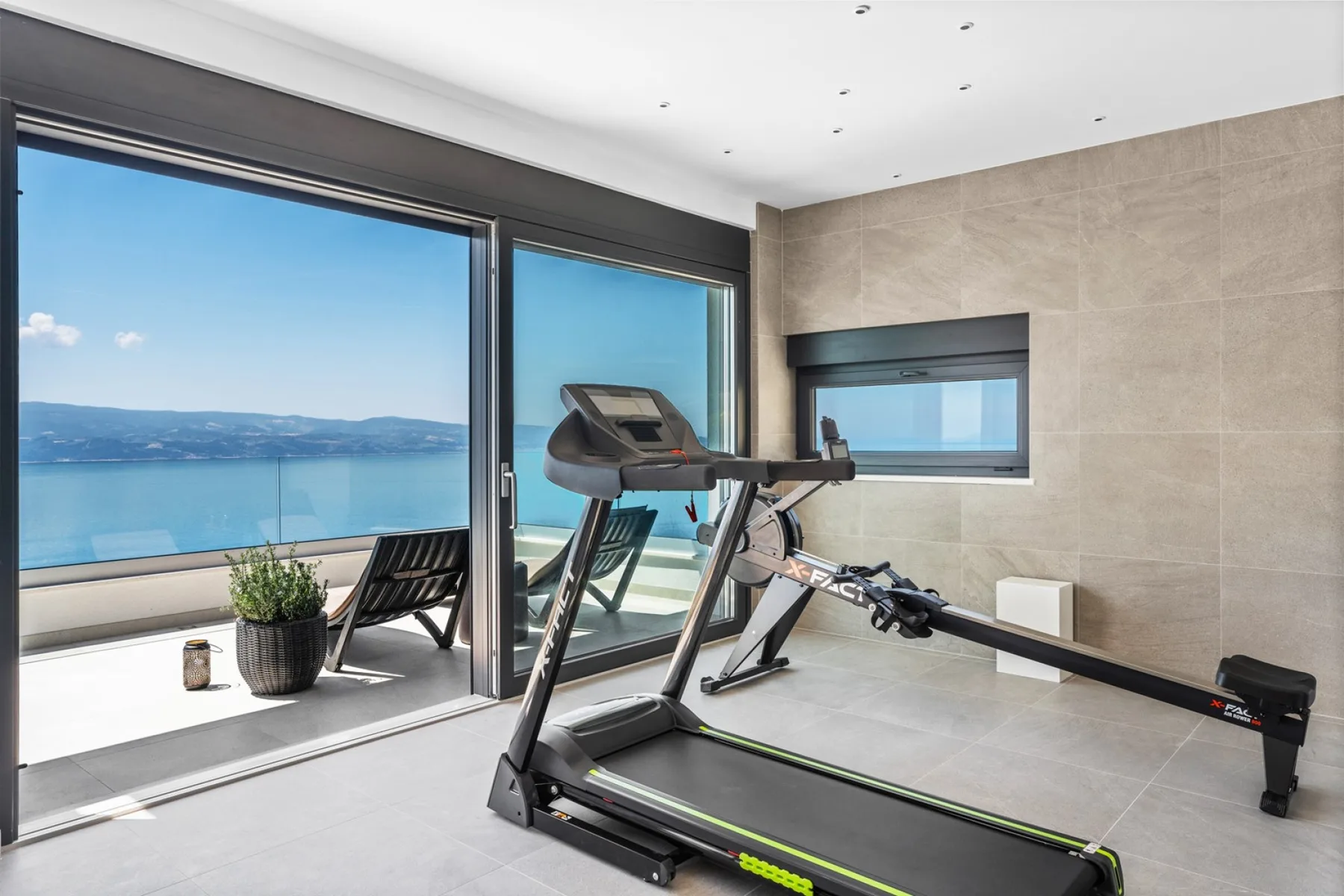A treadmill sits in a bright exercise room beside large windows overlooking the water.