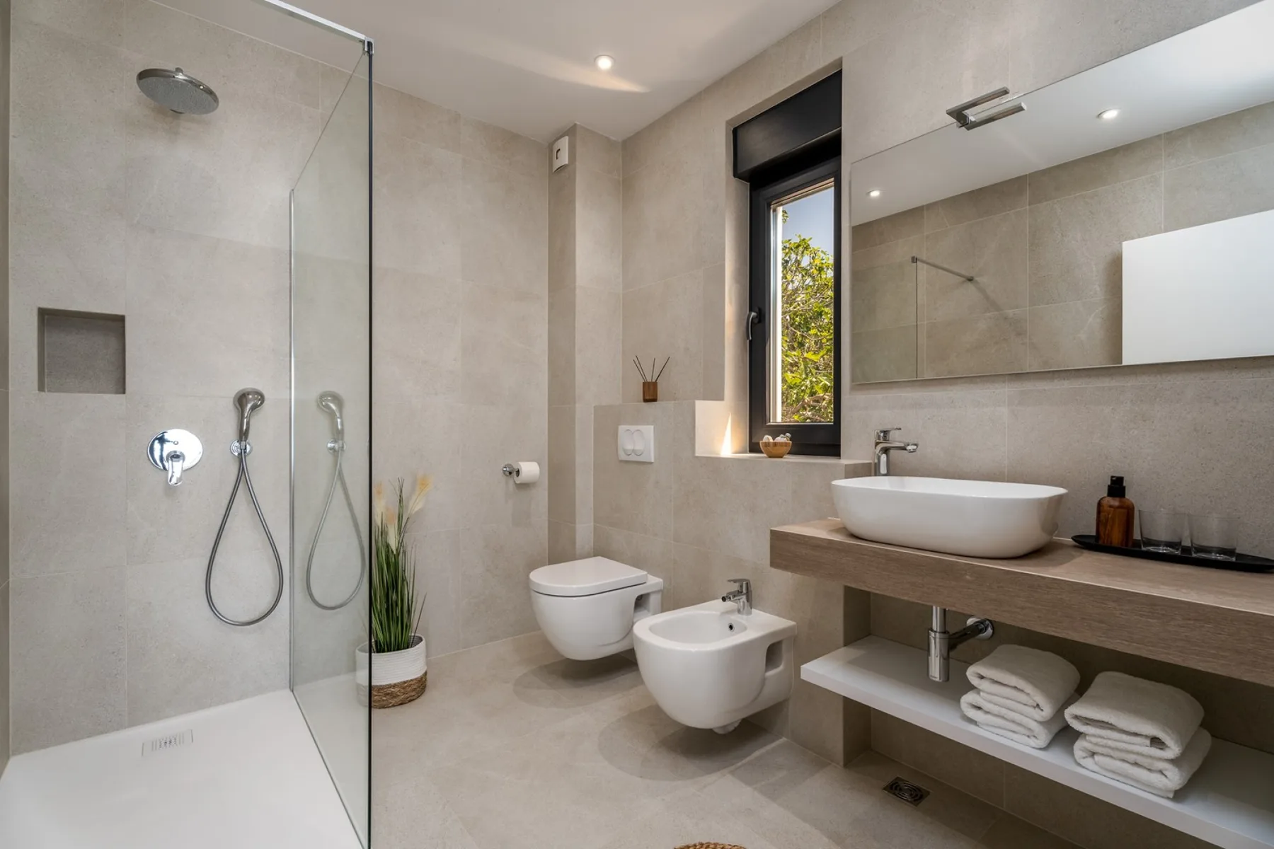A modern bathroom with a glass shower, toilet, bidet, and long vanity.