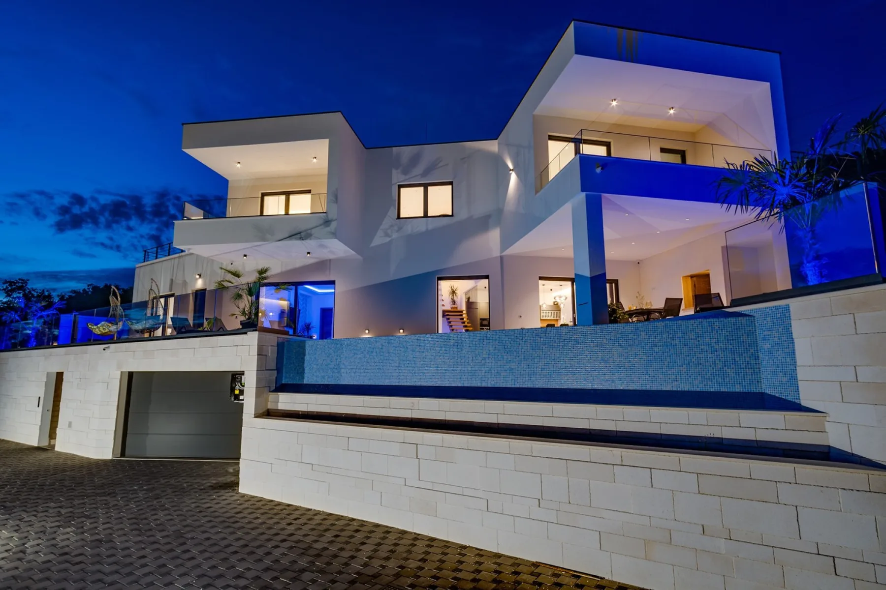 A modern two-story house at dusk, with a blue-lit pool in front.