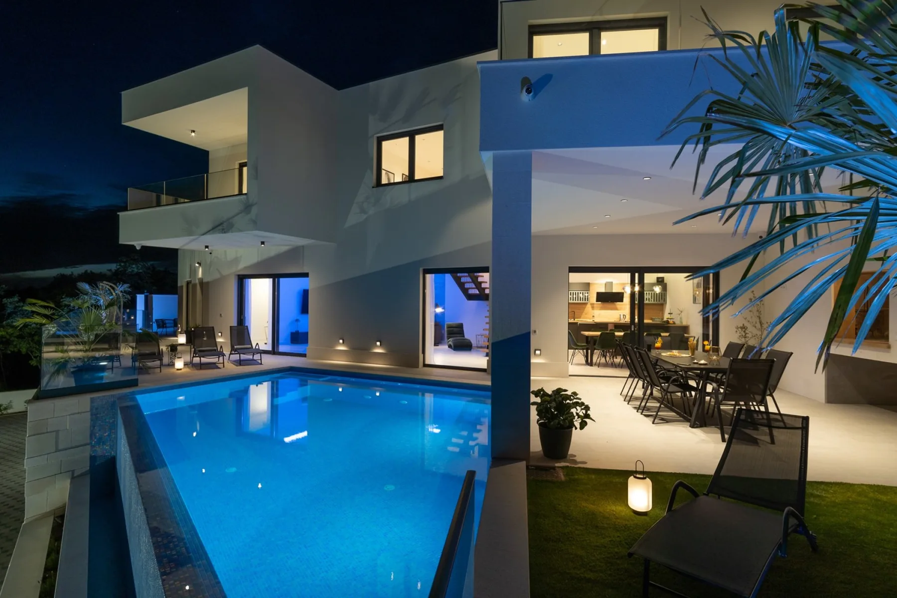 A modern house at night overlooks a blue-lit swimming pool and outdoor dining area.