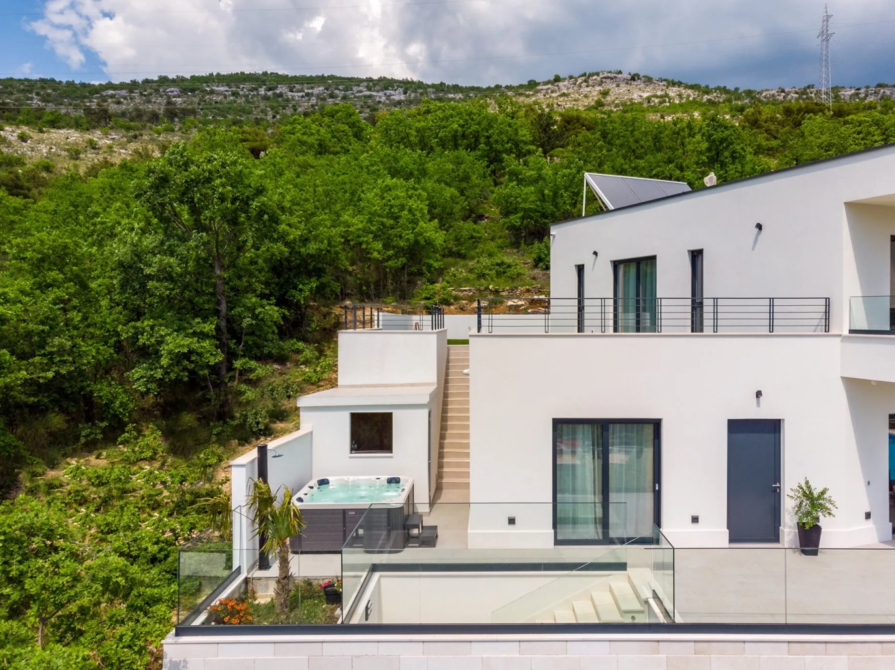 A modern white house with glass-railed terraces sits beside a green hillside.