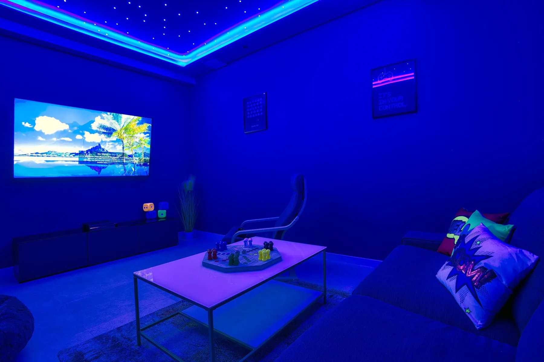 A blue-lit lounge has a wall-mounted screen and a low table in front of seating.