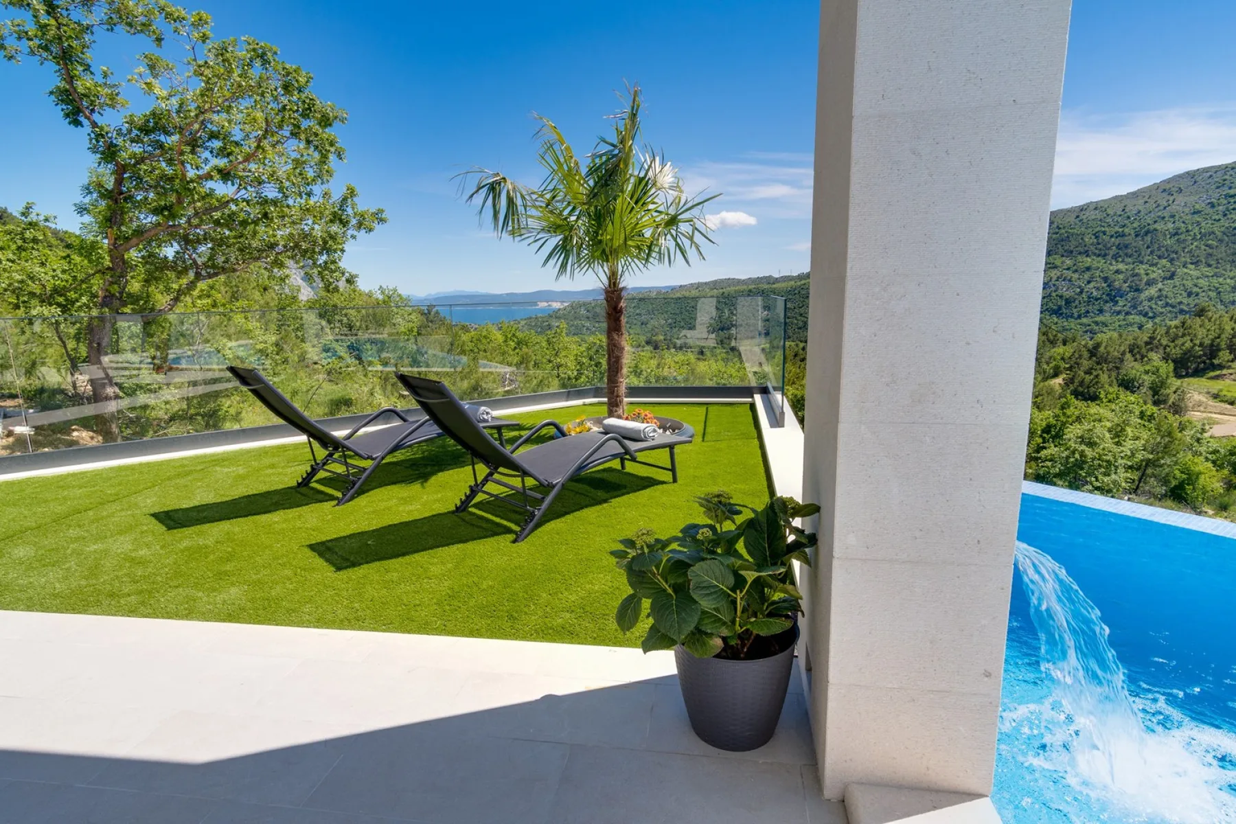 A terrace with lounge chairs, a small palm, and a view of the pool and hills.