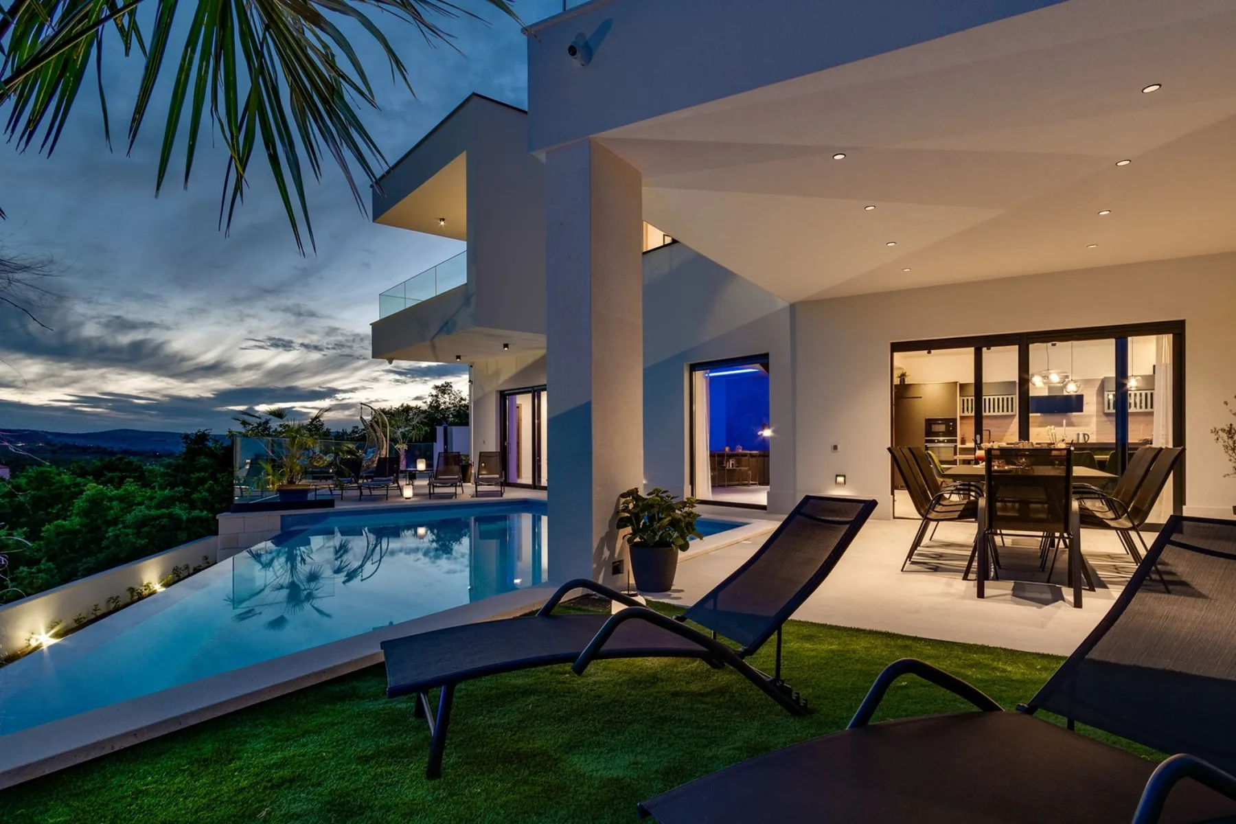 A modern house opens onto a swimming pool and an outdoor dining area at dusk.
