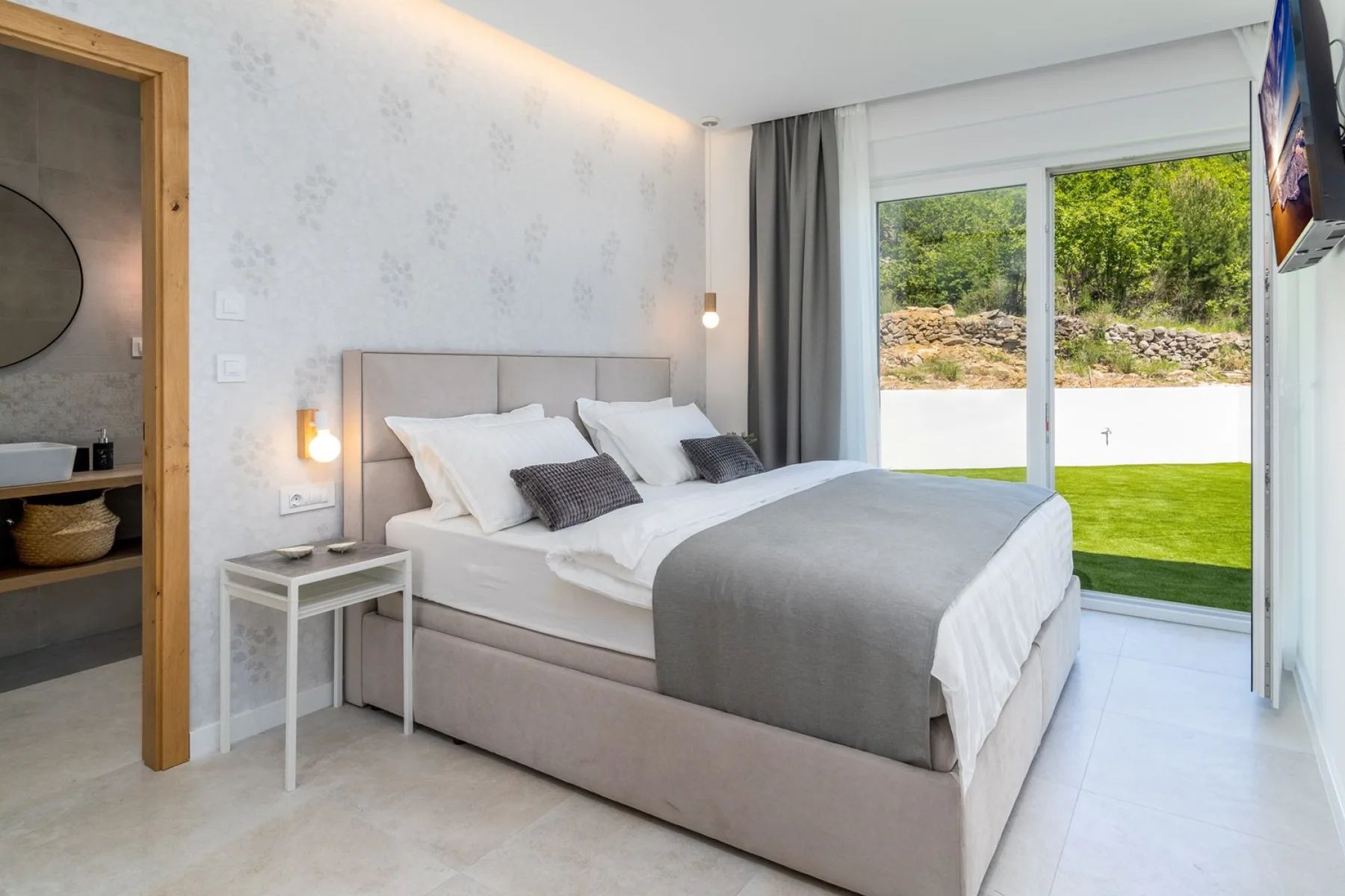 A bright bedroom with a neatly made bed beside large glass doors opening onto a lawn.