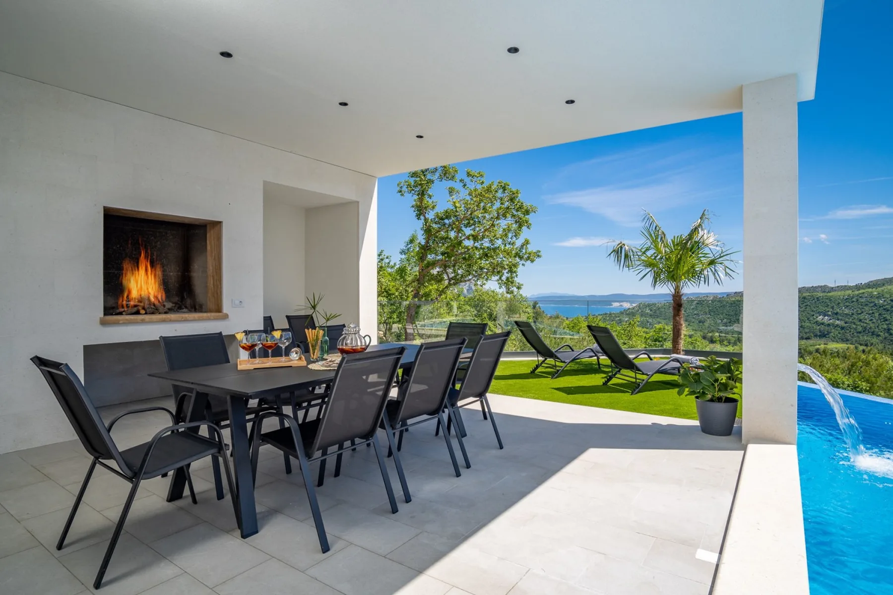 A covered outdoor dining area overlooks a pool, garden, and distant water.