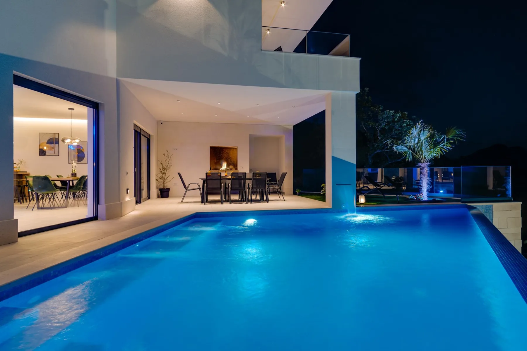A modern home opens onto a lit swimming pool and covered outdoor dining area at night.