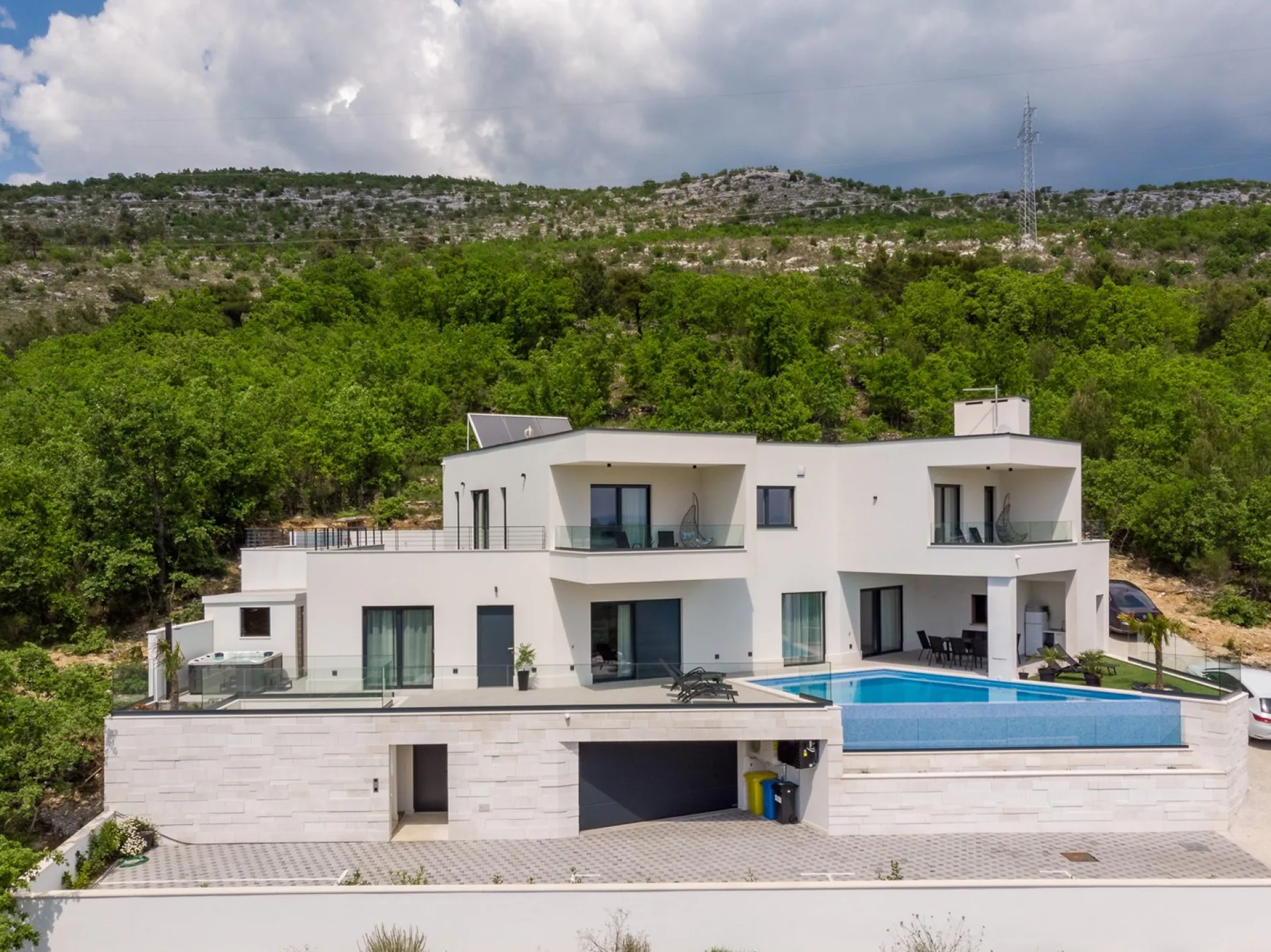 A modern white house with a swimming pool sits beside a green hillside.