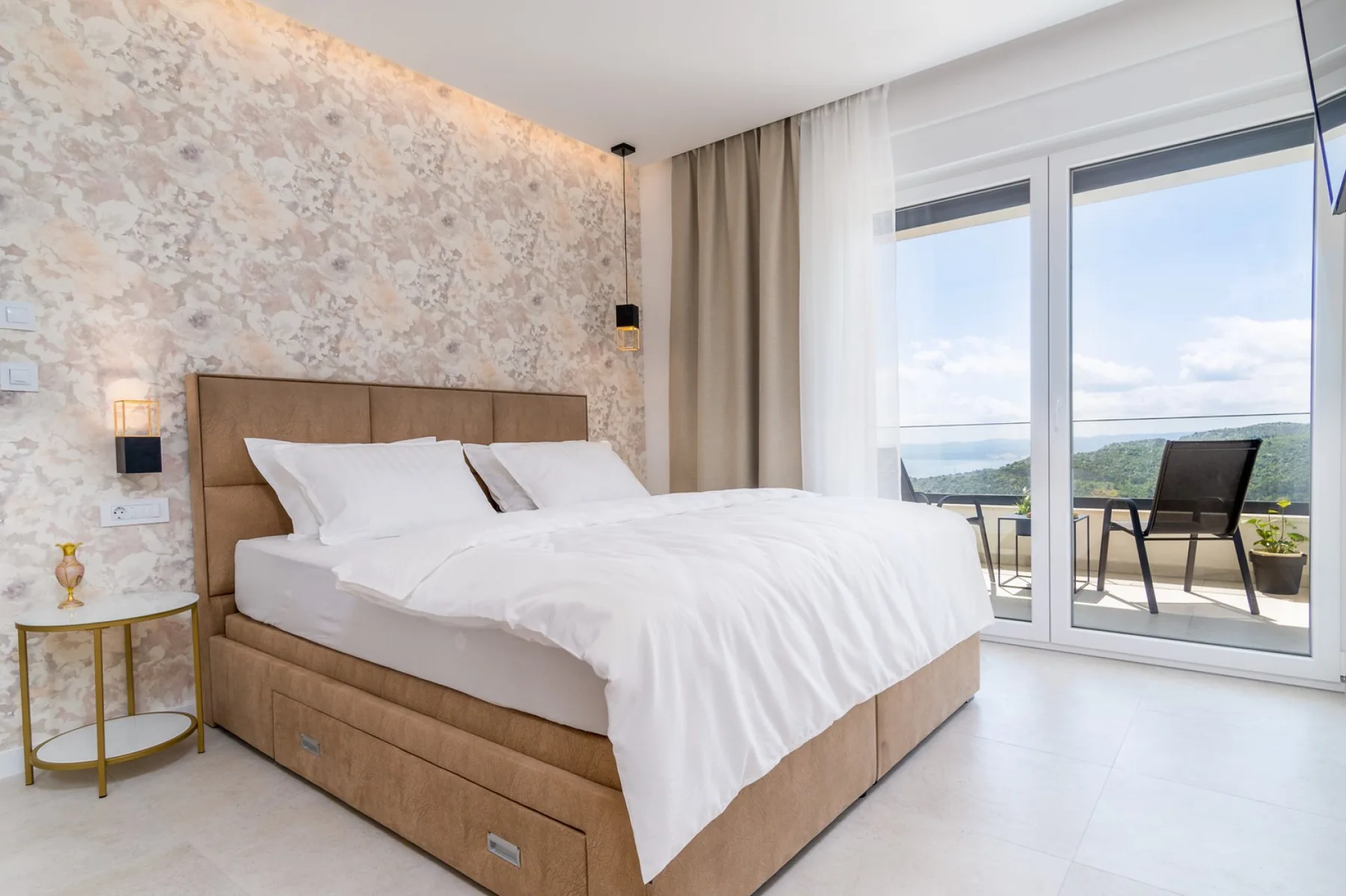 A bedroom with a large bed beside floor-to-ceiling windows opening onto a balcony.