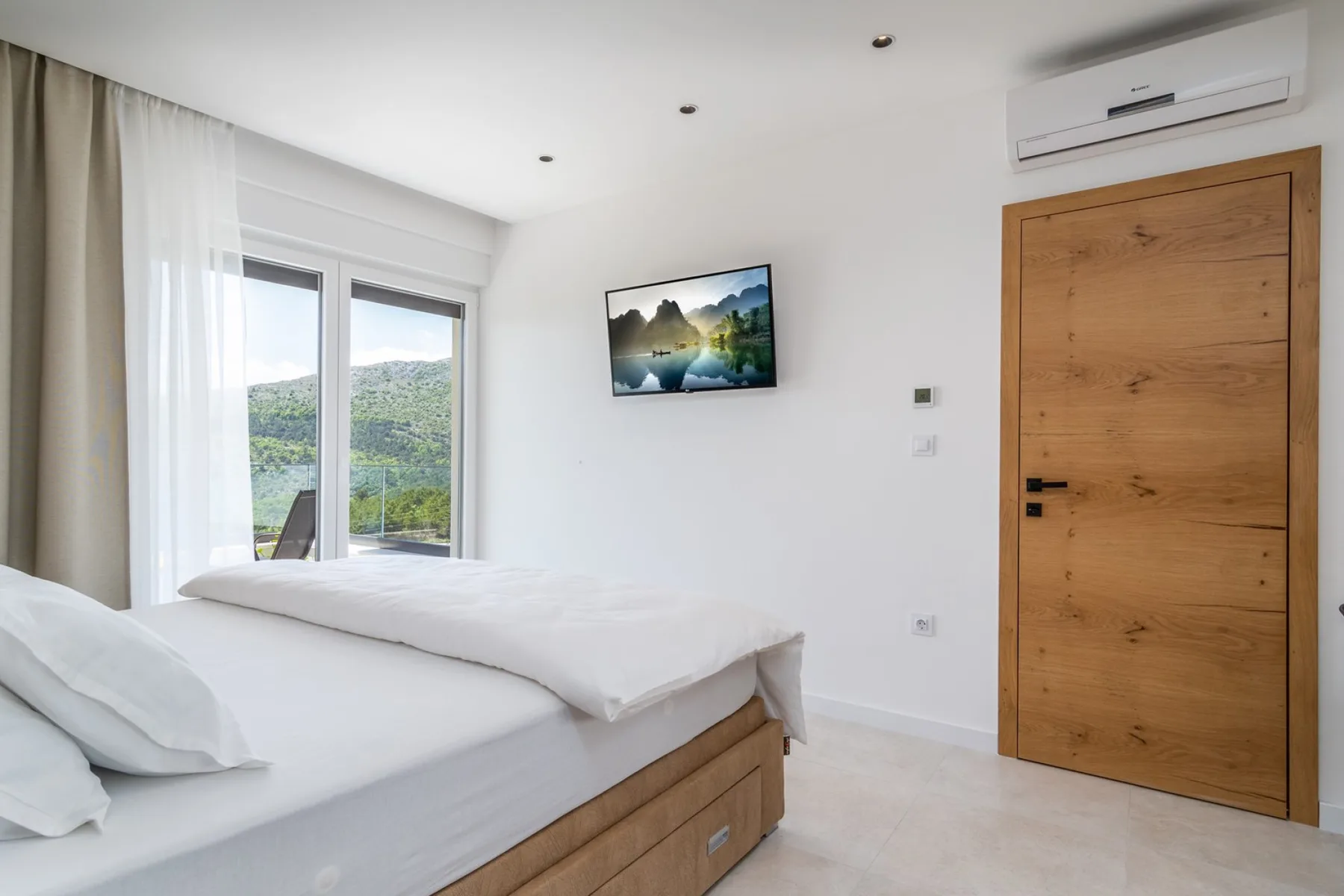 A bedroom with a bed beside tall windows overlooking green hills.