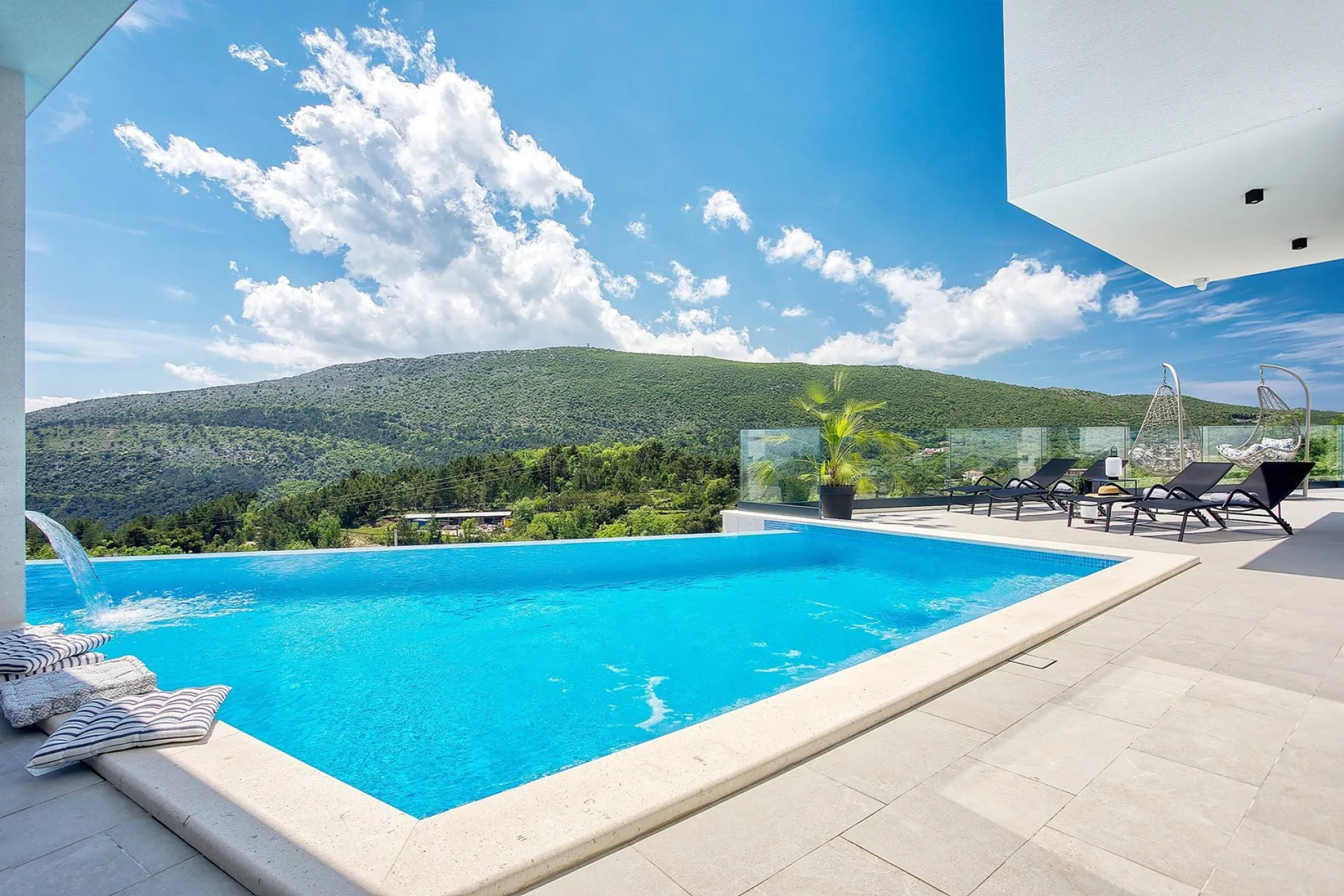 An outdoor swimming pool overlooks green hills beneath a blue sky.