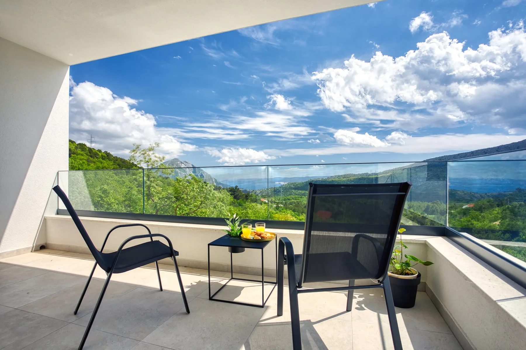 A sunny balcony with outdoor seating and a glass railing overlooking a green landscape.