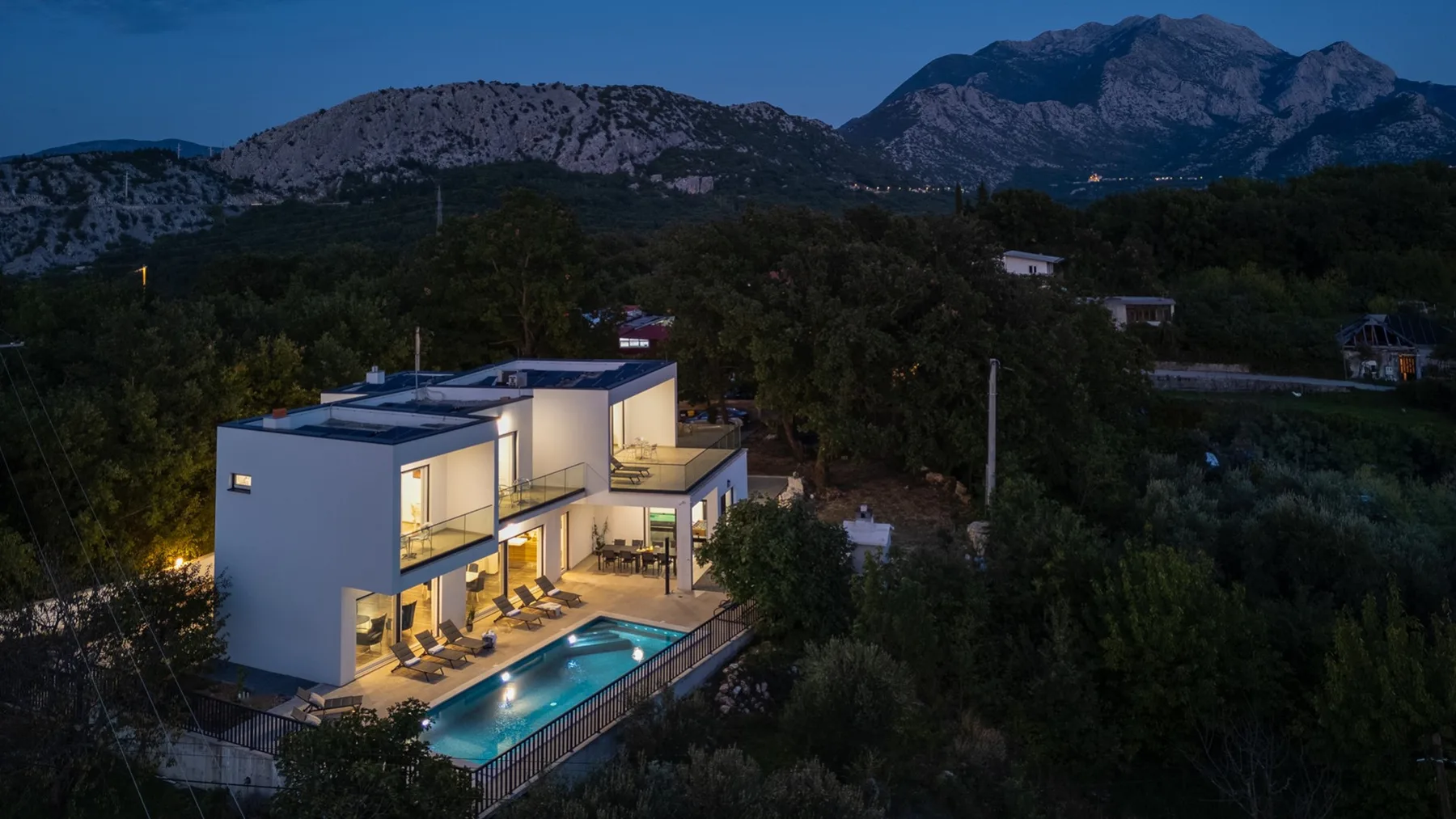 A modern house with a lit swimming pool sits among trees beneath a mountain range at dusk.