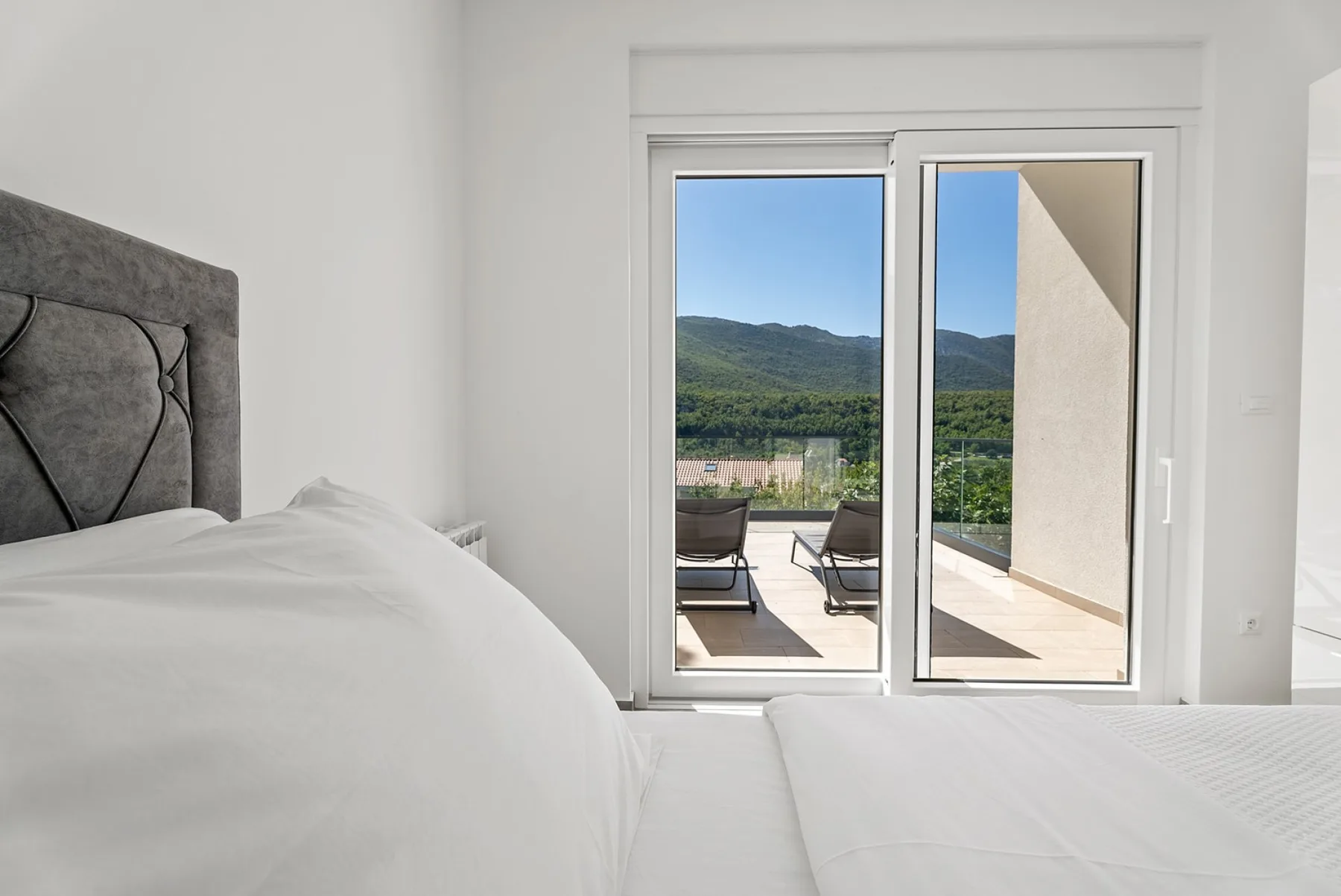 A bright bedroom opens onto a terrace with seating and a mountain view.