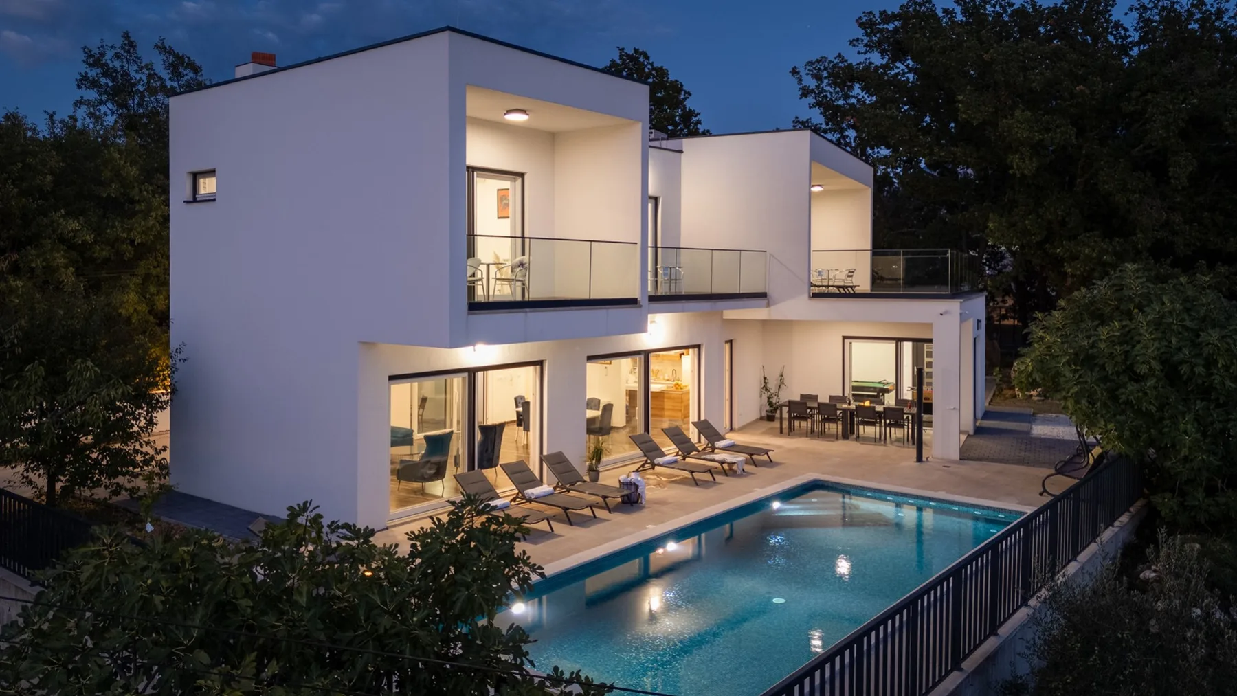 A modern house with balconies overlooks a lit outdoor swimming pool.
