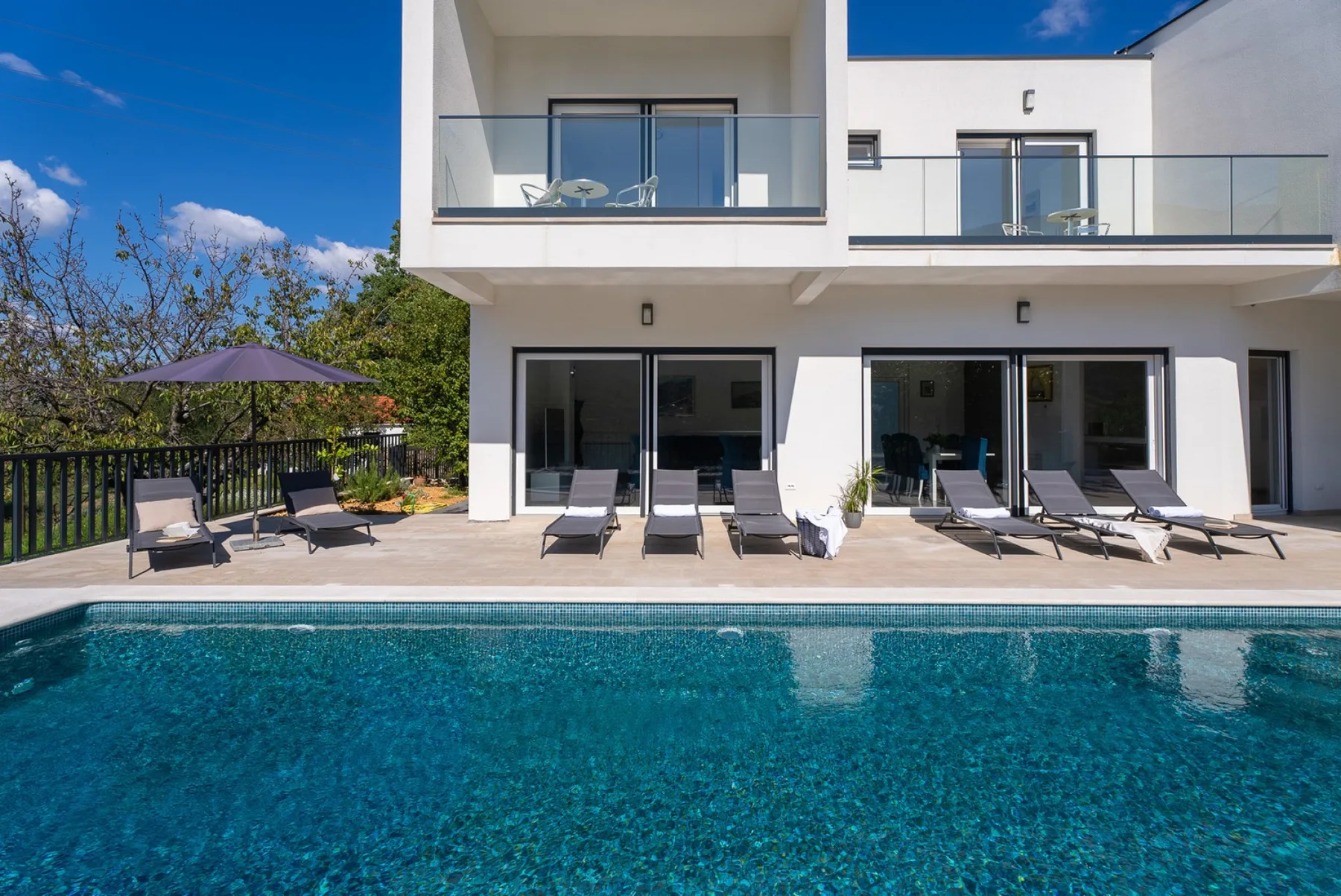 A swimming pool sits in front of a modern white house with patio seating.