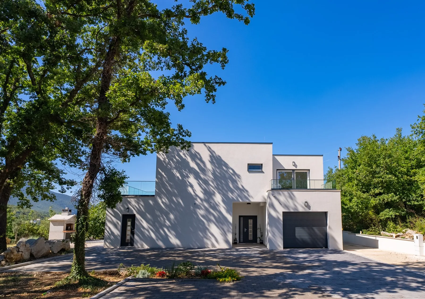 A modern house stands beneath leafy trees beside a paved driveway.