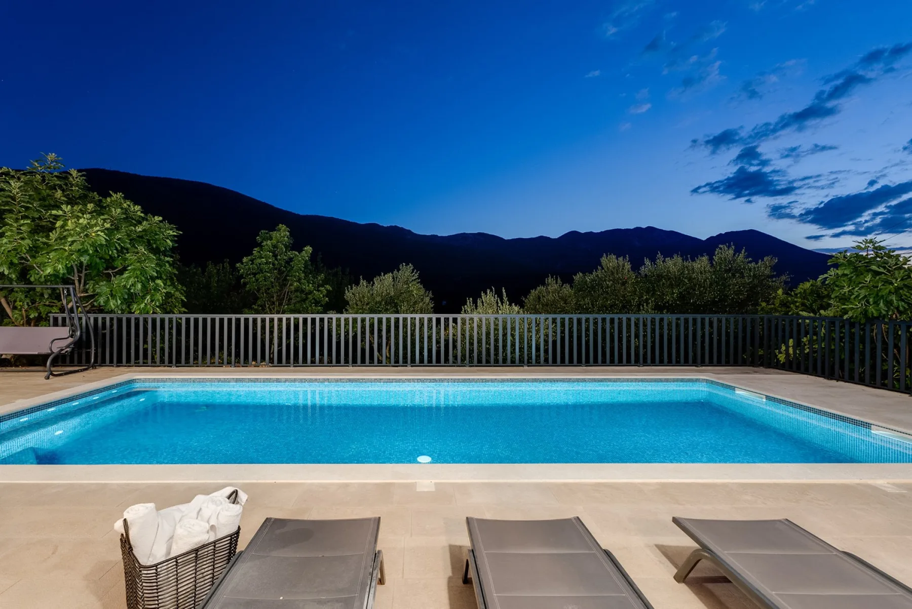 An outdoor swimming pool is bordered by lounge seating and a mountain view at dusk.