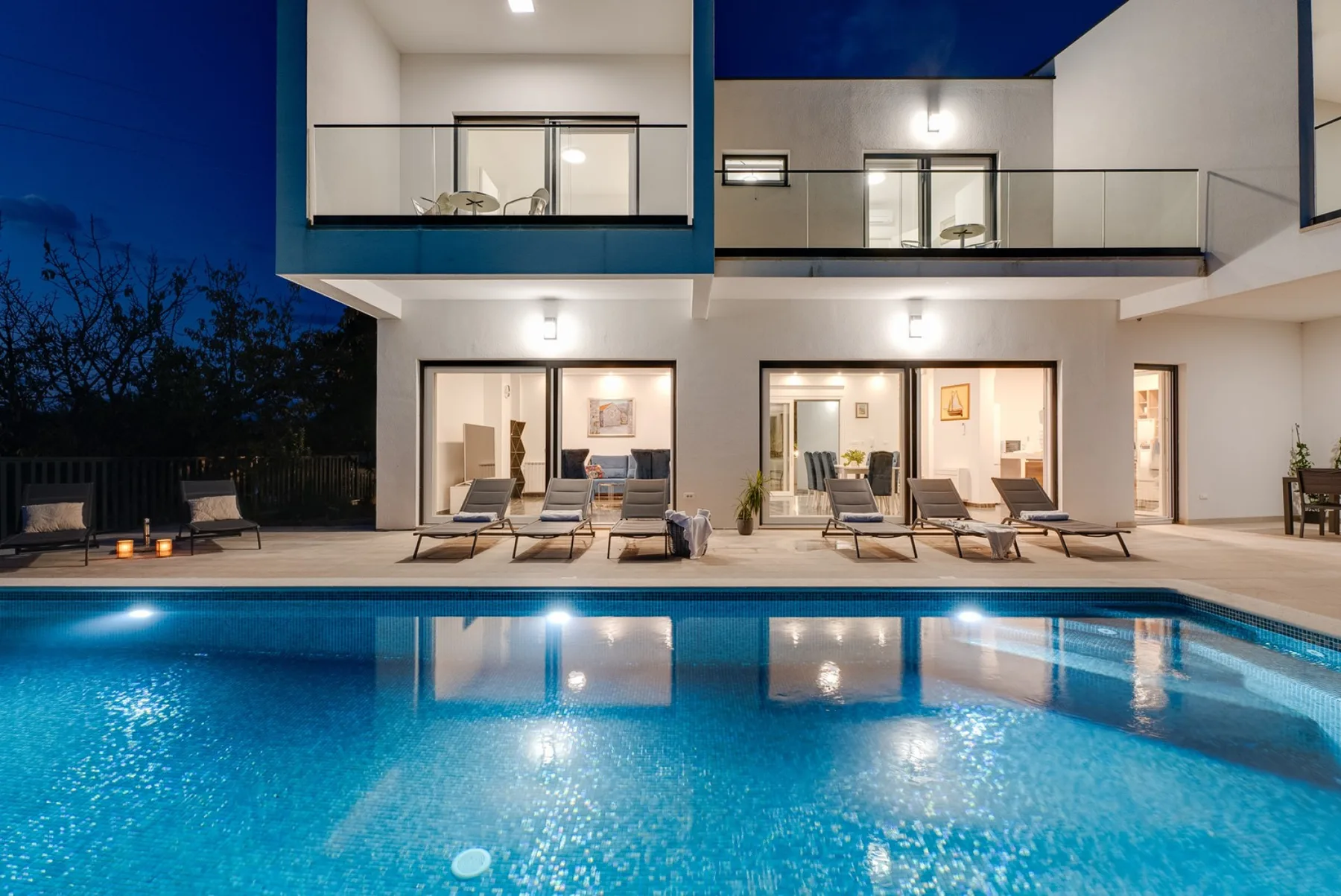 A modern house overlooks a lit swimming pool and outdoor seating at dusk.