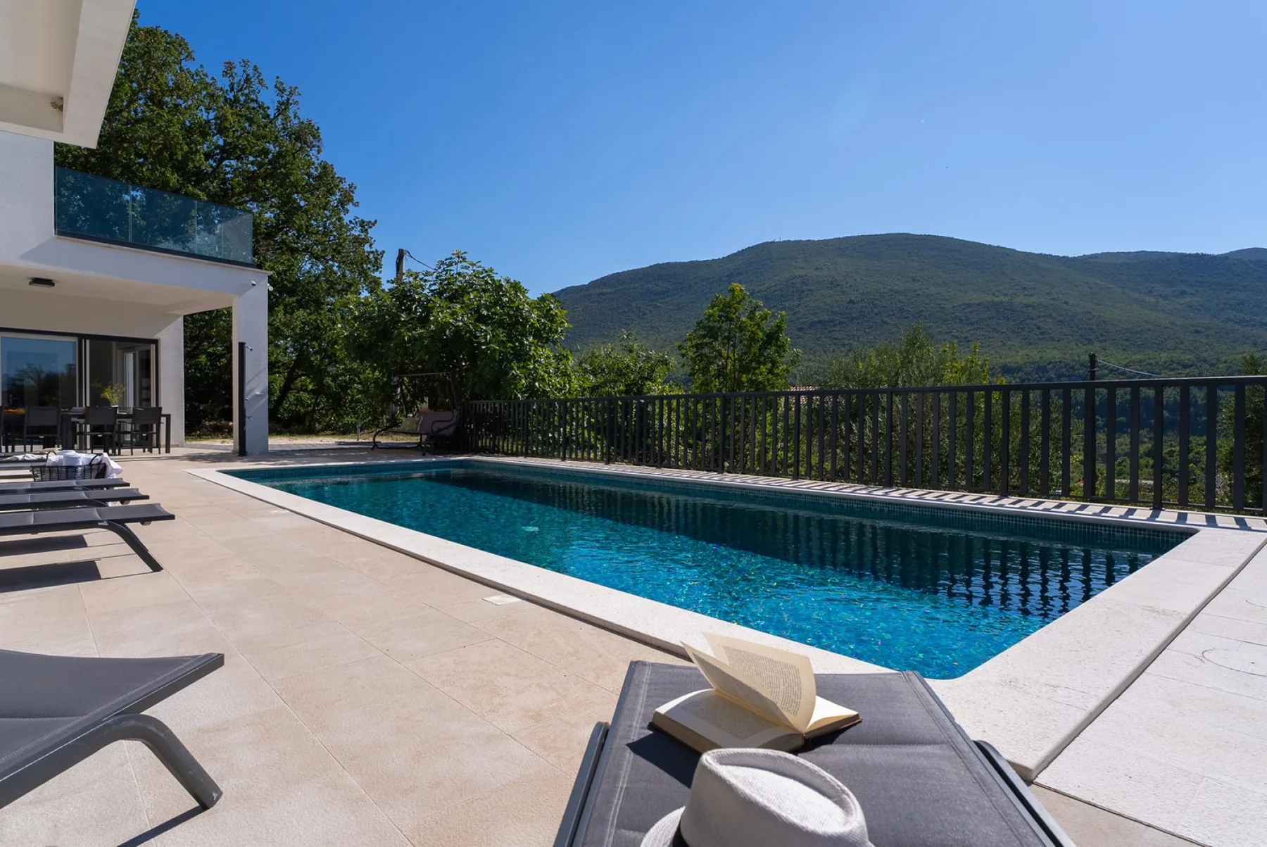 Outdoor pool beside a modern house, with lounge seating and green hills in the distance.